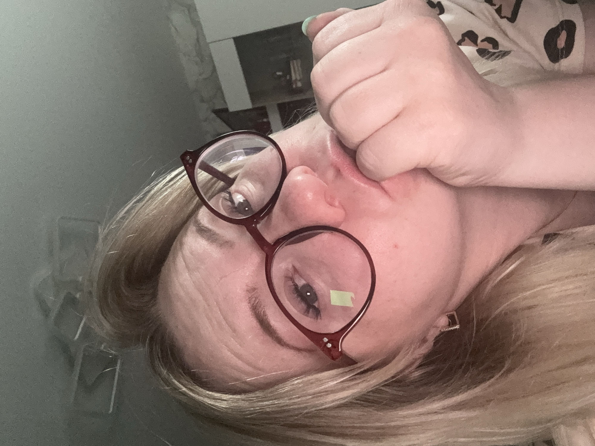 A selfie taken from a low angle depicts a woman with blonde hair wearing red-rimmed glasses. She is holding her fist to her face, near her mouth, with her left hand. A small green sticker is visible on the lens of her glasses over her left eye. She is wearing a patterned shirt. The background is blurry, but some furniture and a light-colored wall are visible. There is no information available from the image that indicates the location to be Chaikovsky, Russia.