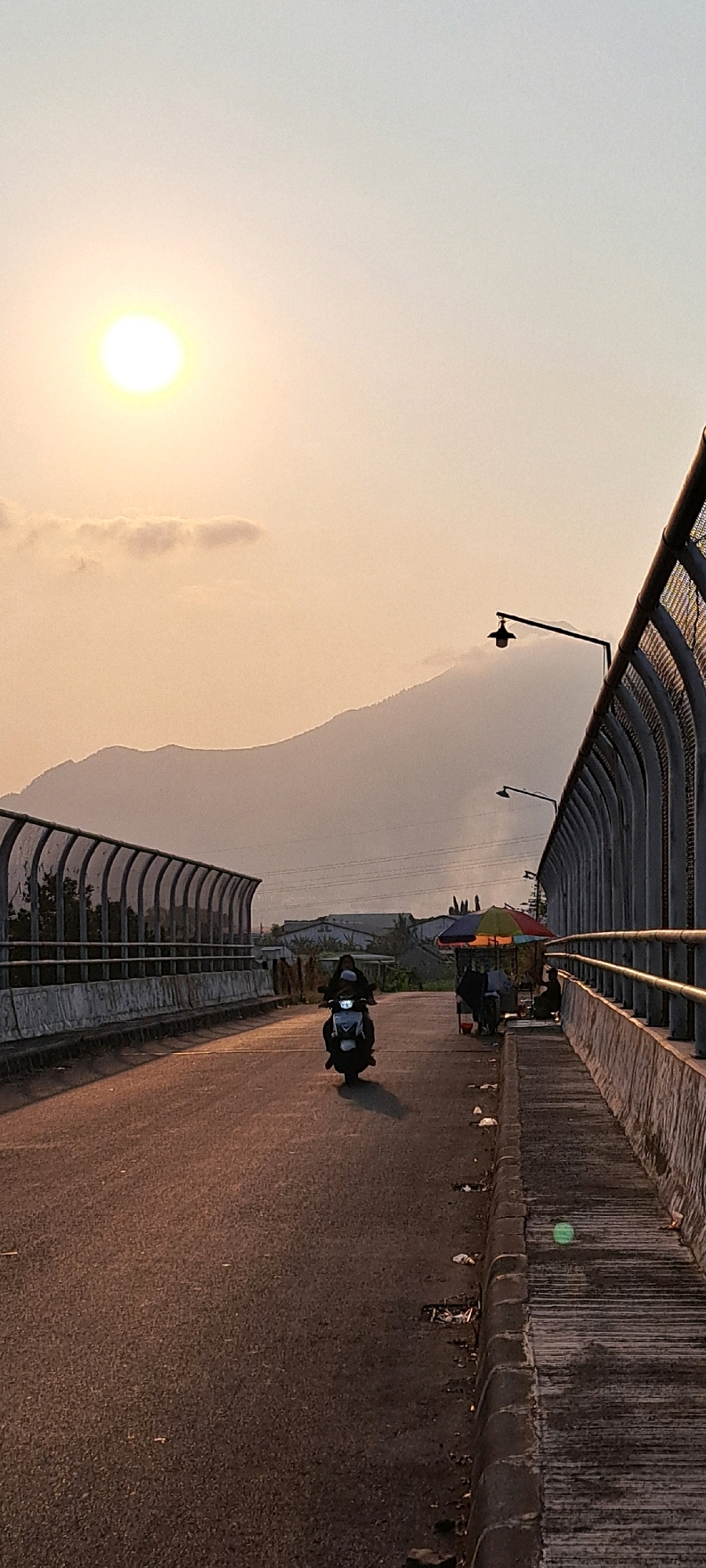 The image depicts a scene on an elevated road, likely a bridge or overpass, during what appears to be sunset. The sun, a bright, diffused orb, is visible in the upper left quadrant, casting a warm, golden light over the hazy sky and the distant mountains. The road surface is asphalt, showing signs of wear and scattered debris along the edges.
On the road, a single person is riding a white scooter, heading towards the viewer. They are wearing a dark helmet and clothing. To the right of the road, a sidewalk or walkway is present, bordered by a metal fence with vertical slats and a top railing. Further down the walkway, near the edge of the frame, a colorful umbrella is set up, under which several people are gathered, suggesting a small roadside stall or resting spot. Two streetlights with distinct lamp housings are mounted on poles along the road's edge.
The environment includes concrete barriers and metal fencing lining both sides of the elevated road. In the background, beyond the road and fences, there are indications of urban or industrial structures and the silhouette of a mountain range. The overall atmosphere is serene, enhanced by the warm lighting and the muted colors of the landscape. The location is identified as Kebon Waris, Indonesia.