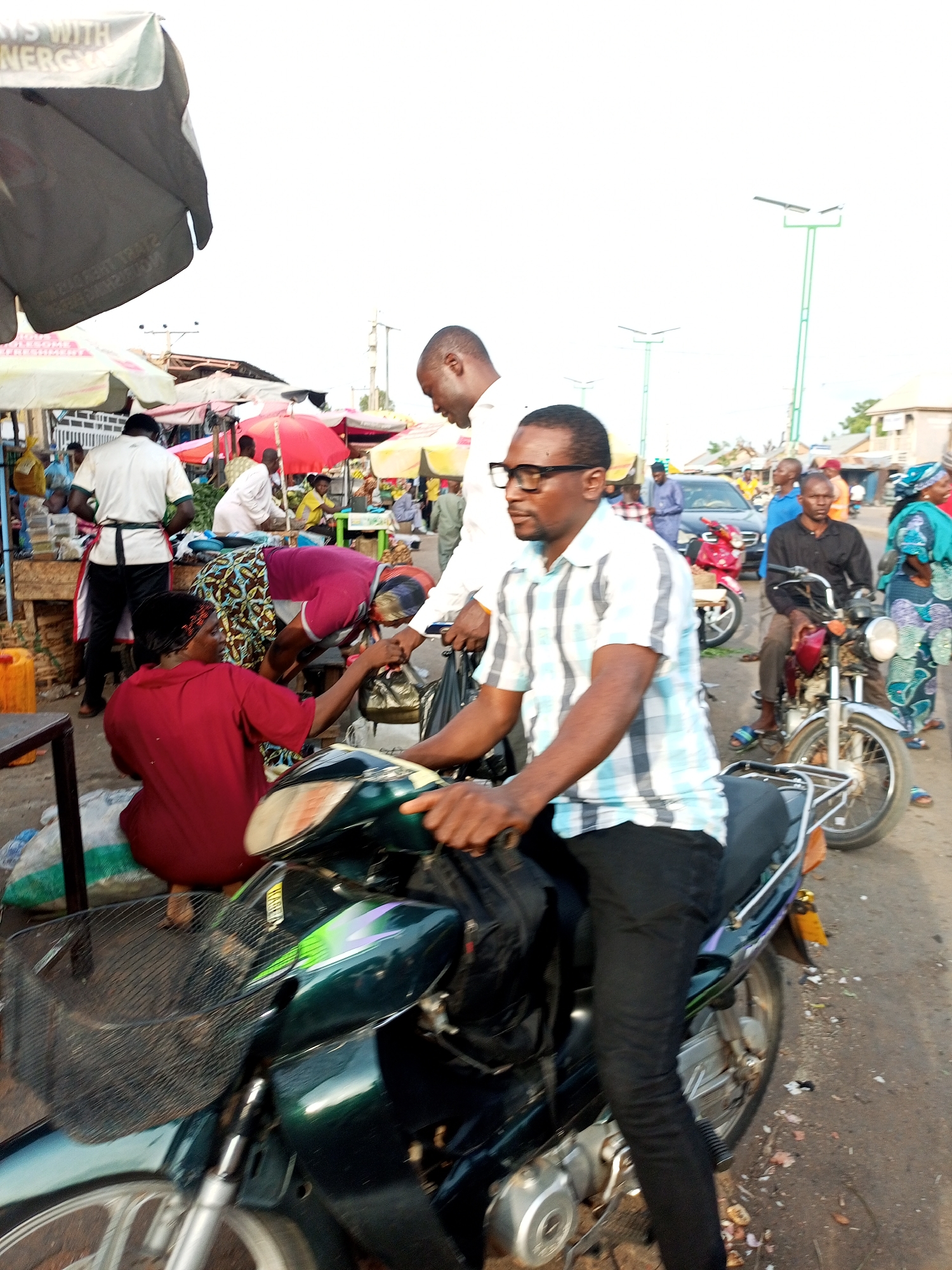 In the city of Tempure, Nigeria, a bustling outdoor market scene unfolds. In the foreground, a man wearing a checkered shirt and dark pants is seated on a dark green motorcycle, looking to the left. He appears to be holding the handlebars. Behind him, a woman in a red shirt is bending over, possibly interacting with the man on the motorcycle or another person. Her hair is adorned with a dark patterned headband.
Further back, another man in a white shirt and dark pants stands near a market stall, his back to the camera. To his right, several other individuals are visible, some standing and some seated. A red umbrella is open at one of the stalls. In the background, more market stalls with various umbrellas are set up, and there are a few motorcycles and a car. The environment suggests a typical day in a busy Nigerian market with open-air stalls and ongoing commerce. The sky is overcast and bright.
