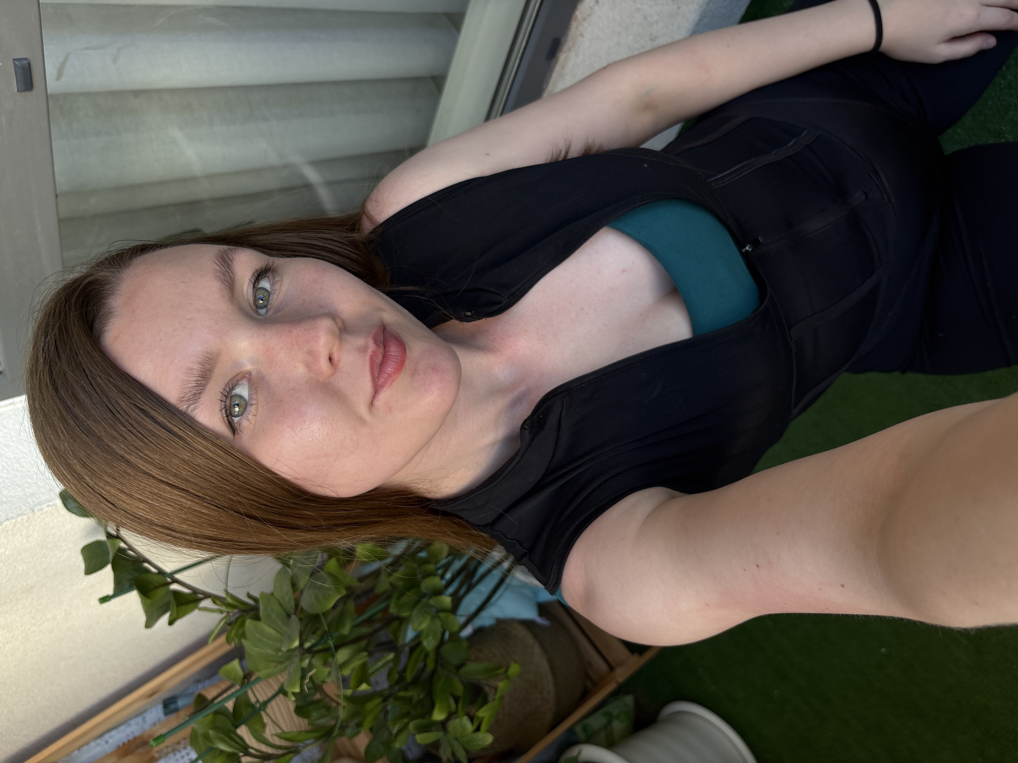 A young woman with brown hair and green eyes is taking a selfie on a balcony. She is wearing a black sleeveless vest over a teal sports bra and black leggings. Behind her, there is a potted plant and a window with vertical blinds. The balcony has a green turf floor. The overall setting suggests an outdoor urban environment.