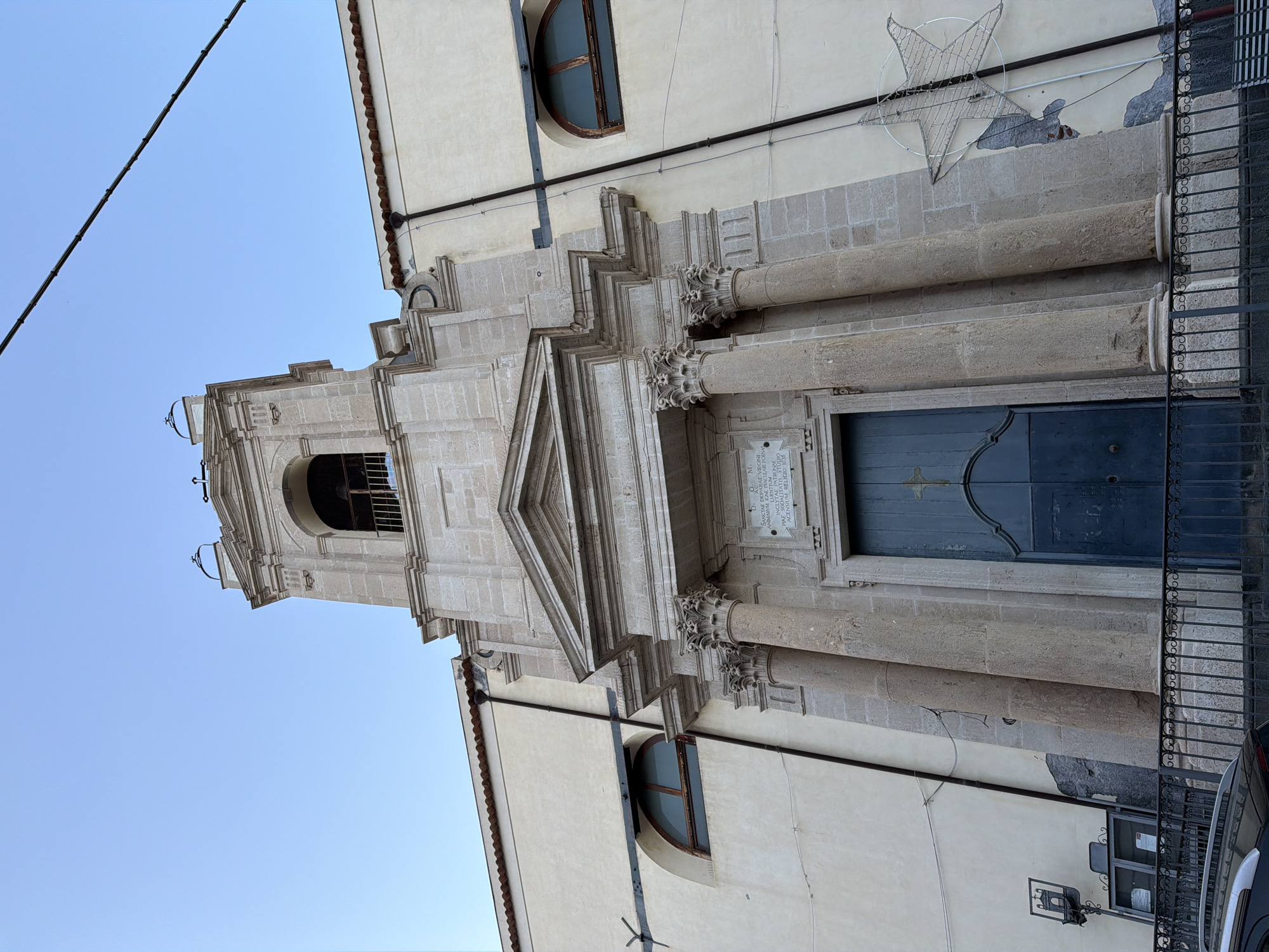 Here is a detailed description of the image:
The image captures an angled view of a building facade in Acireale, Italy. The architectural style suggests a historical or religious structure, likely a church or similar edifice.
The facade is characterized by elaborate stonework, including decorative columns with intricate capitals, a triangular pediment above the entrance, and a square bell tower with an arched opening. A blue door is prominent and features an embossed cross. A small plaque mounted on the stone facade contains Latin text.
To the right of the entrance is a metal fence. A lit star hanging from the roof is visible.
The sky is visible above the building, indicating daytime. A black power cable is prominent on the left side. The building has windows with rounded tops.