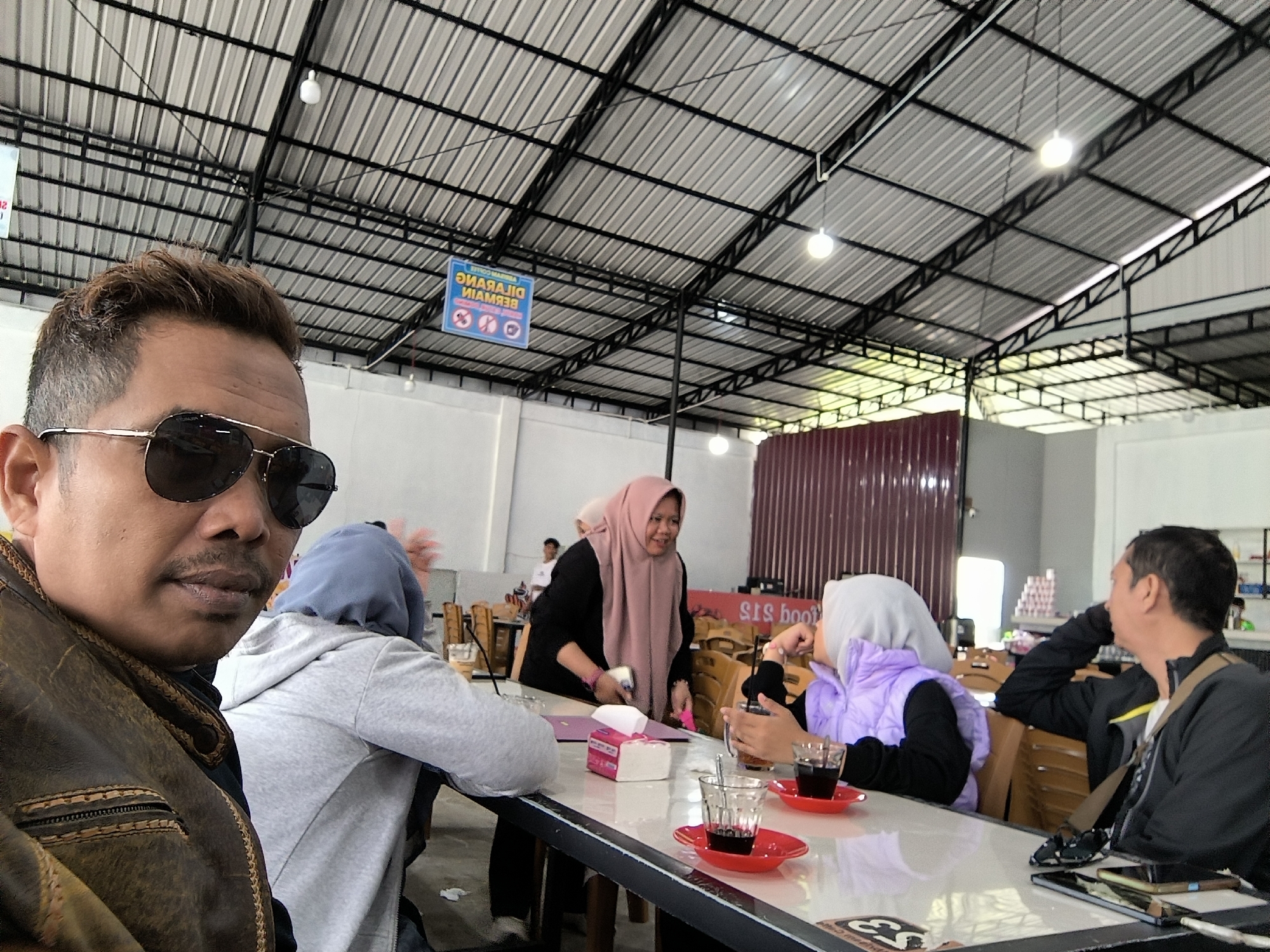 This media file depicts a group of people gathered at a table in what appears to be an outdoor or semi-outdoor dining area, likely a cafe or restaurant in Panteraya, Indonesia. The setting features a corrugated metal roof supported by a black metal framework, with industrial-style lighting fixtures. The overall atmosphere suggests a casual daytime gathering.
In the foreground, a man wearing sunglasses and a brown leather jacket is looking towards the camera, taking up a significant portion of the frame. He has a mustache and short, styled hair. Behind him, several individuals are seated around a table. One woman in a pink hijab is smiling and gesturing while holding a smartphone and a pink box of tissues. Another person wearing a light blue hijab and a grey hoodie is also seated at the table. Across from them, a woman in a purple vest and hijab is holding a drink, and a man in a black jacket is looking away, resting his head on his hand.
On the table are several items, including glasses with dark liquid (likely coffee or tea), red saucers, straws, a pink tissue box, and what looks like a smartphone. The table itself is white and appears to be a laminate surface.
In the background, there are other tables and chairs, suggesting a public dining space. A maroon-colored wall is visible on the right. There is also a sign hanging from the ceiling with Indonesian text, partially obscured but appearing to be a warning or informational sign with symbols. One visible sign has text that reads "DILARANG MEROKOK" which translates to "NO SMOKING." The lighting suggests it is daytime. The mood appears relaxed and social.