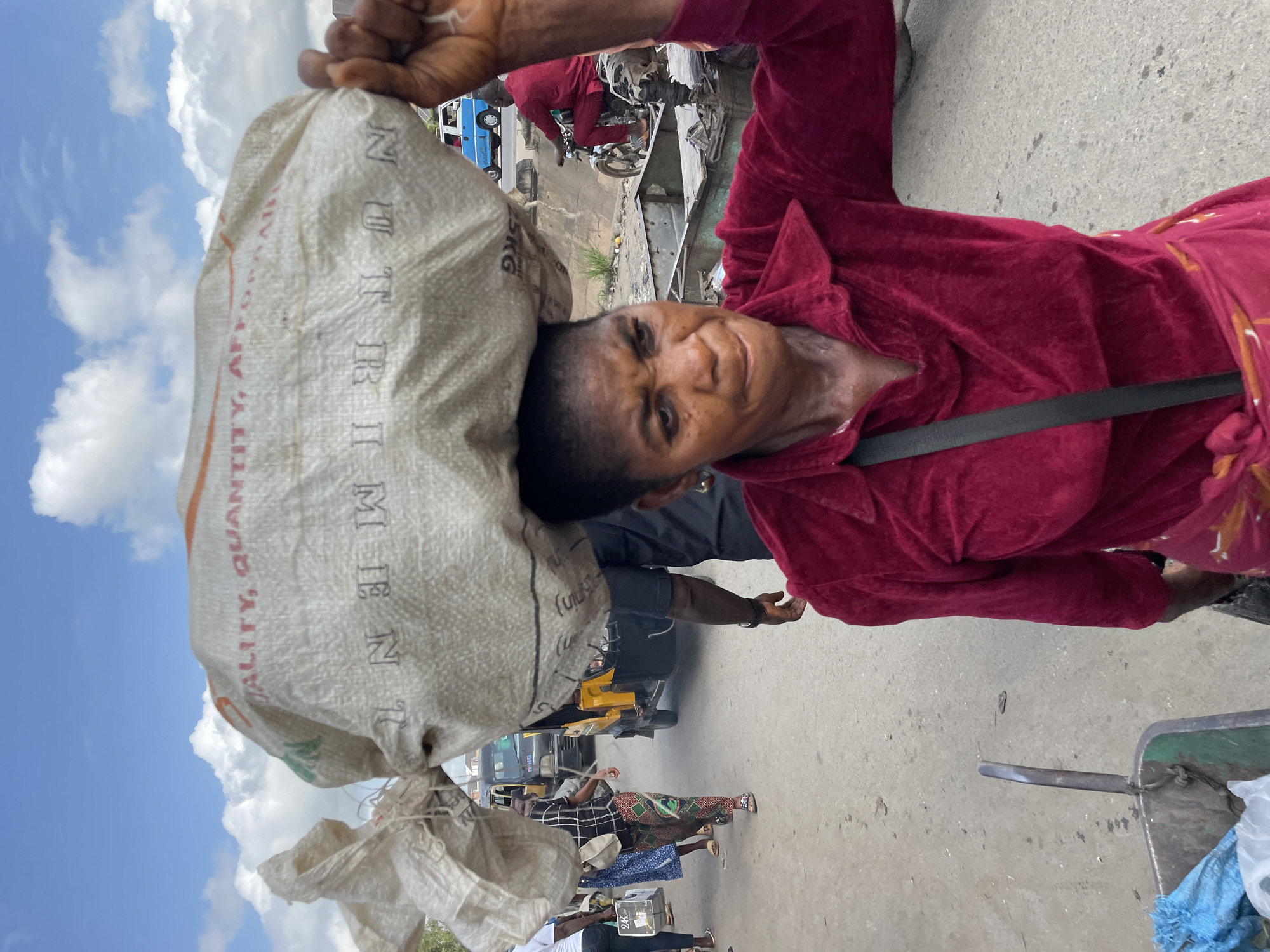 A woman in Fegge, Nigeria, carries a large sack on her head. She is wearing a red velour robe, and her face is turned towards the camera. Her expression appears to be one of strain or exertion. The sack is made of woven material and has some text on it, though it is partially obscured. Visible words on the sack include "QUALITY, QUANTITY" and "MEN".
The scene appears to be outdoors, with a cloudy sky in the background and a paved surface underfoot. Other people and vehicles, including a blue bus and a yellow auto-rickshaw, are visible in the background, suggesting a market or busy street environment. The time of day appears to be daytime, and the weather seems overcast. The image has a vertical orientation.