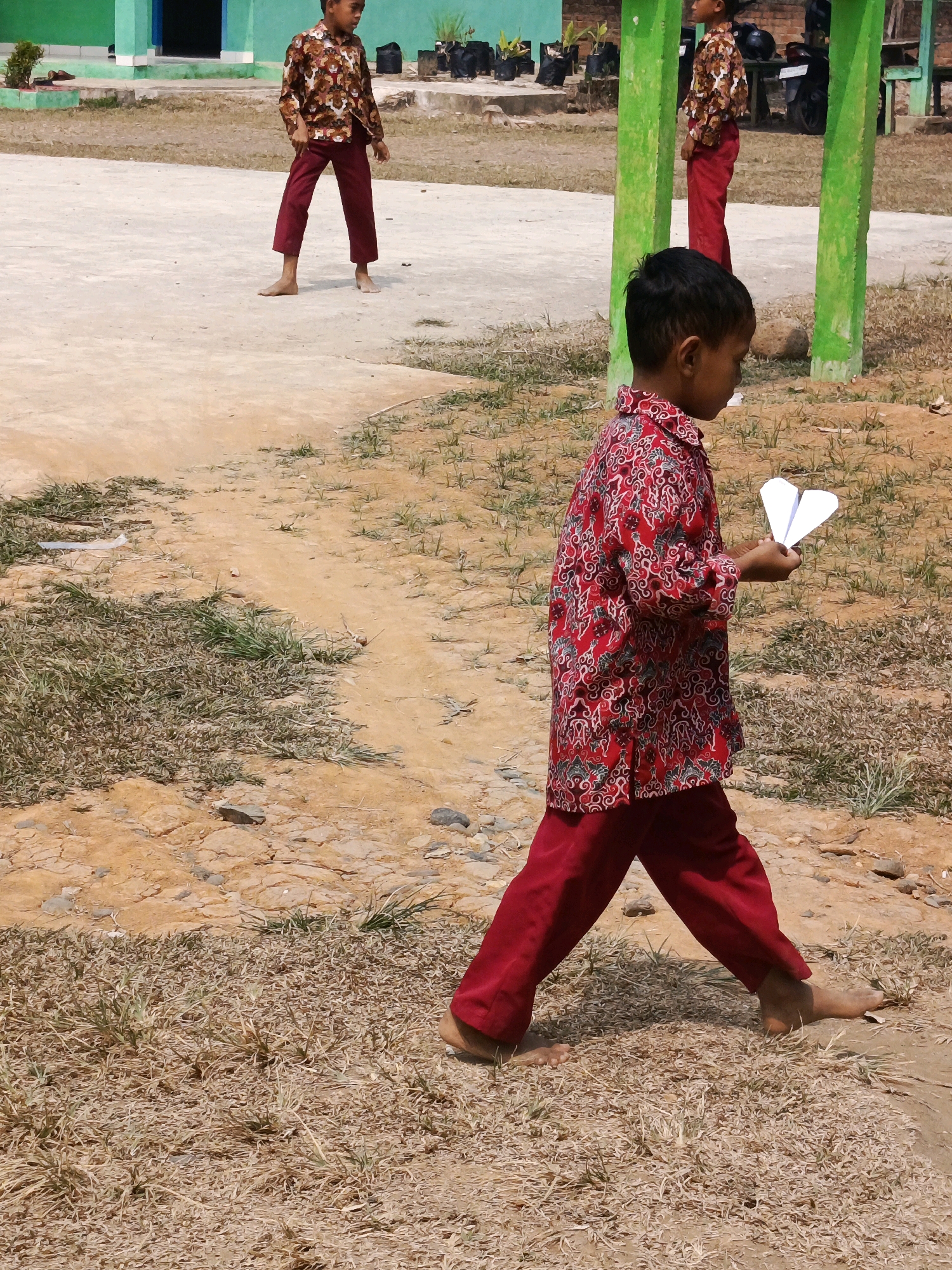 In the outdoor area of Megang Sakti IV, Indonesia, a young boy walks towards the right of the frame, holding a white paper airplane in his right hand. He is wearing a red long-sleeved shirt with a white and green patterned design and matching red pants. He is barefoot. Behind him, another boy, also barefoot, stands on a concrete surface, wearing a similar patterned shirt and red pants. In the background, a light green building with a dark entrance is visible. The ground is a mixture of dirt, sand, and dry grass. There are green posts and black trash bags scattered in the background.