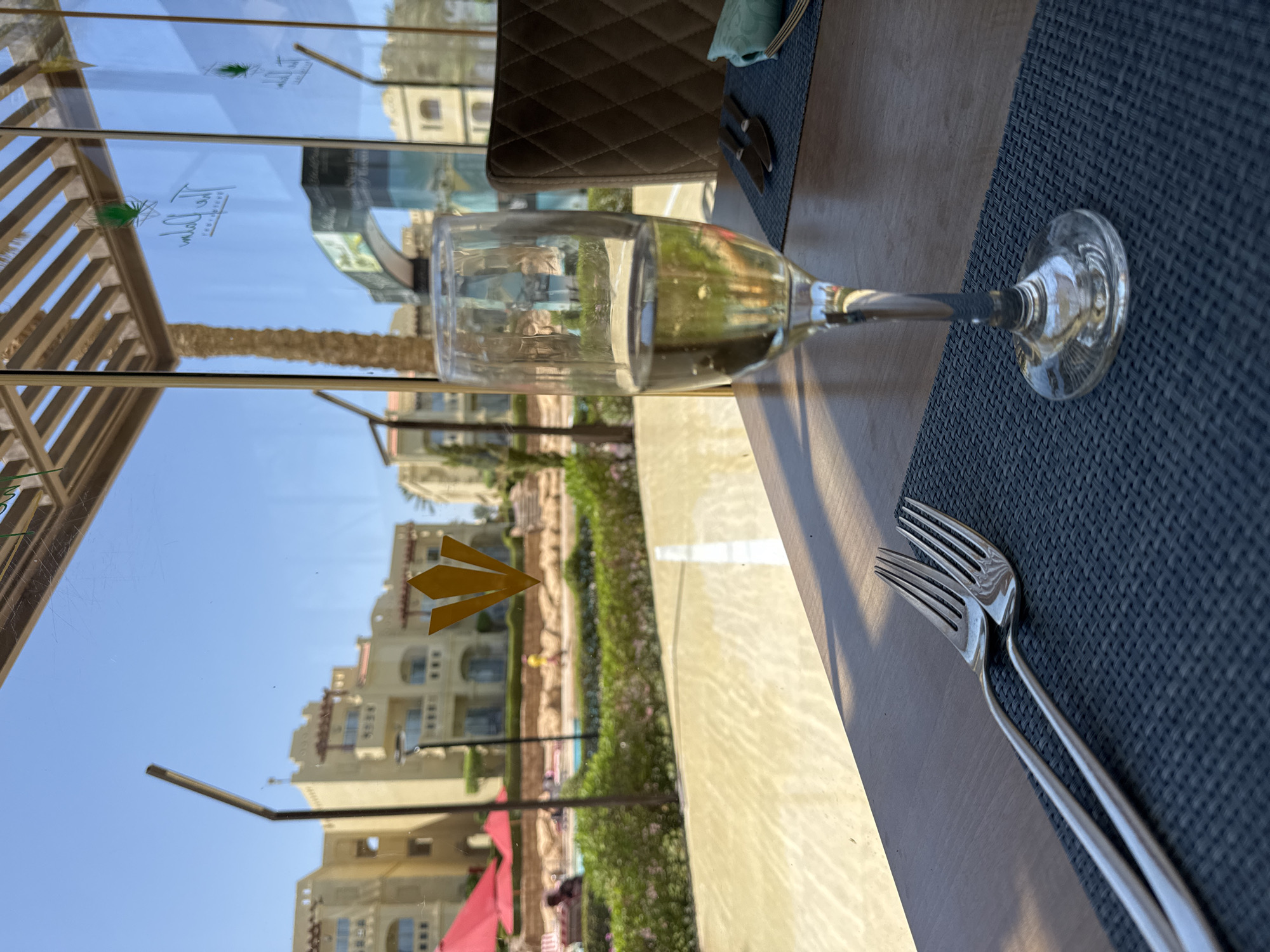A close-up shot focuses on a glass of white wine on a wooden table, with two forks placed to the left on a dark placemat. The background, viewed through a glass window, reveals a sunny outdoor scene. The view includes a swimming pool with red umbrellas, lush green bushes, and buildings with a sandy, stucco exterior, characteristic of a resort in an Egyptian location. The window also displays reflections of the interior, including a logo with a palm tree. The overall ambiance suggests a leisurely setting at a resort.