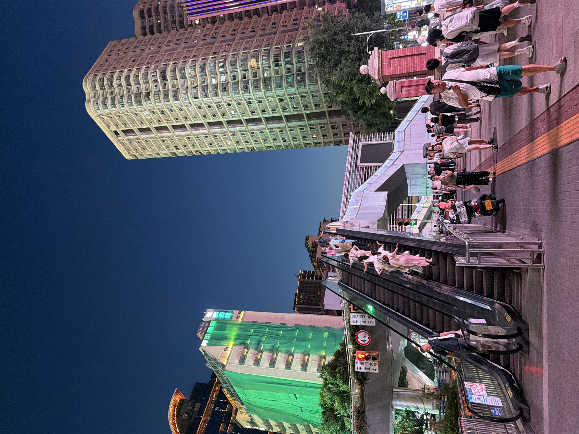 In the Yuzhong District of China, this scene unfolds at dusk, featuring a vibrant cityscape with modern architecture and bustling pedestrian activity. Dominating the right side of the frame is a tall, light-colored residential building characterized by its curved balconies and numerous windows, illuminated by interior lights. Adjacent to it, another similar building is partially visible. On the left, a multi-story building is partially obscured by a large, bright green tarp, suggesting ongoing construction or renovation. A prominent escalator runs diagonally upwards, populated by a group of individuals dressed in light-colored, flowy attire, appearing to ascend. Below the escalator, a man in a dark shirt and pants is descending, carrying a white bag. The street level is paved and illuminated, with several pedestrians walking and standing. Traffic lights are visible, including a red signal. A motorcycle is parked on the right side of the sidewalk. The sky is a deep, clear blue, transitioning from twilight into night.