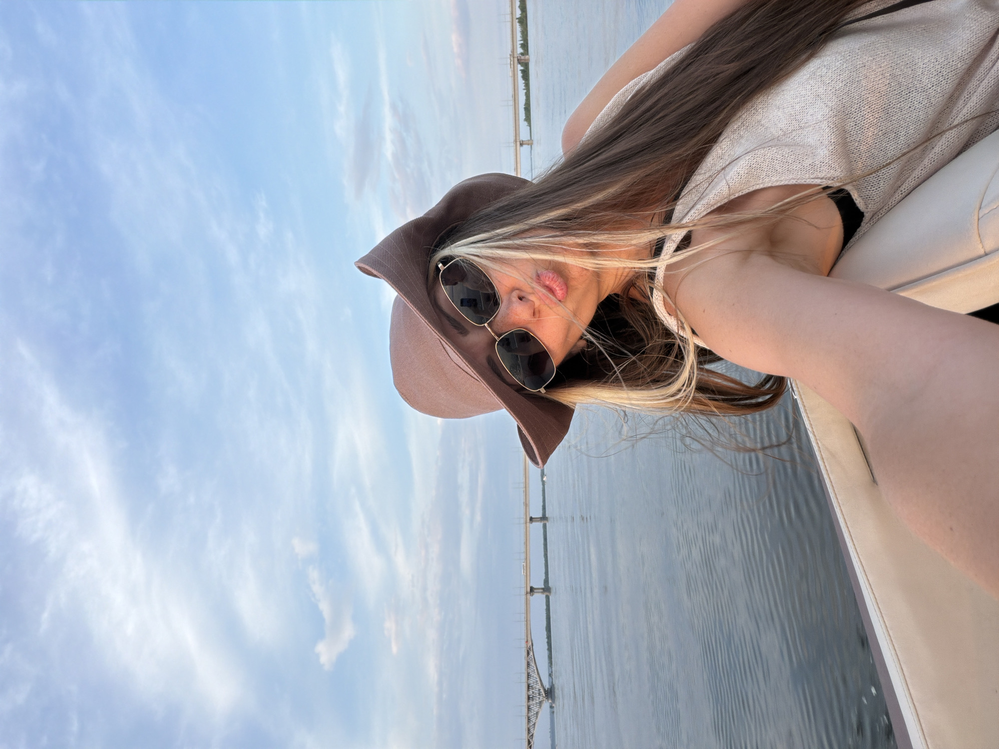 A woman with long, light-brown hair and blonde highlights, wearing a brown hat and sunglasses, is taking a selfie while on a boat on the Volga River. She is puckering her lips. In the background, a long bridge spans the river. The sky is a pale blue with scattered clouds. The location is the Volga River near Saratov, Russia.