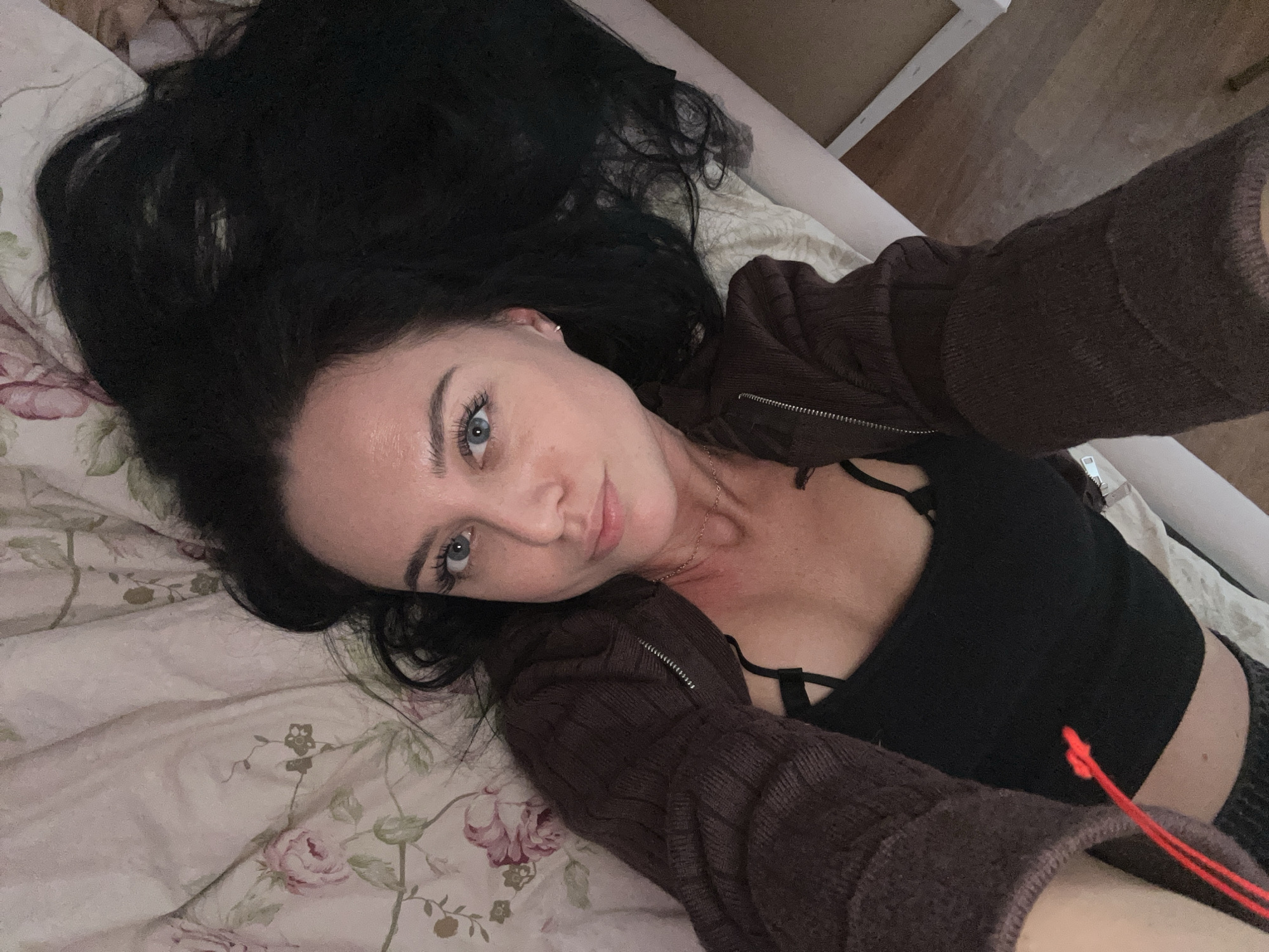 A woman is lying on a bed with floral patterned sheets, taking a selfie. She has long, dark hair spread out on the pillow. She is wearing a black top and a brown zip-up sweater. Her skin appears fair, and she has light blue eyes. The bed is near a white wall and possibly a wooden floor. There is no indication of the location being Chaikovsky, Russia, in the image.