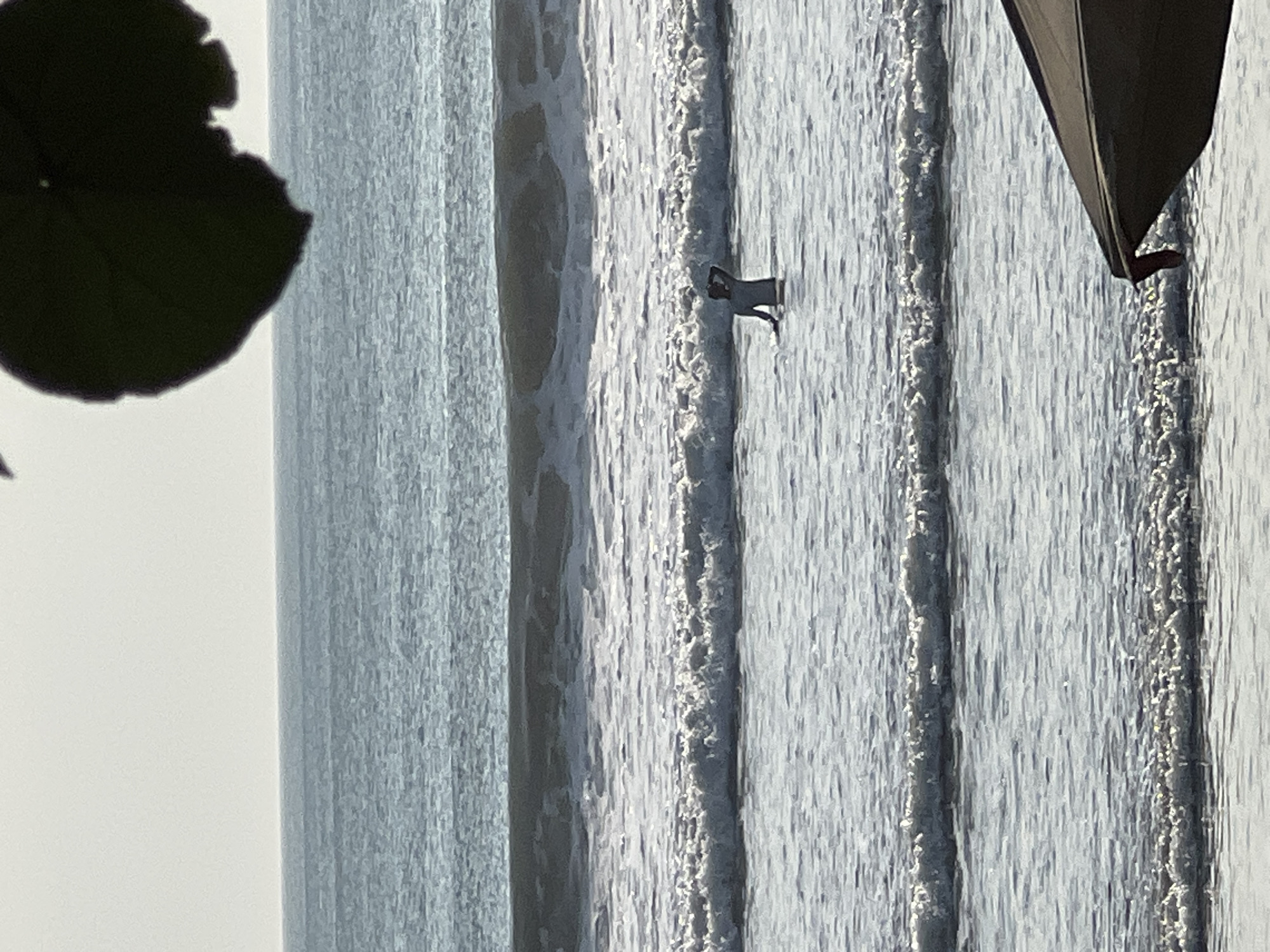 The image shows a person in the ocean in Kuta, Indonesia. The person is standing in the water, which is about waist-deep. They are wearing a light-colored long-sleeved shirt and dark pants. The person is facing away from the camera and appears to be looking out at the ocean. There are several waves in the ocean, and the water is choppy. The sky is overcast, and the light is muted. In the foreground, part of a dark-colored umbrella is visible, suggesting the photo was taken from a beach.