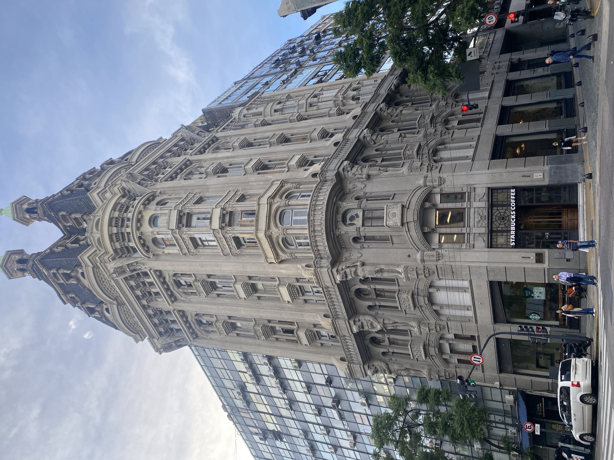 The image shows the Palacio Bencich in Buenos Aires, Argentina, an ornate, grey stone building with intricate facade details. A modern glass building is adjacent to the Palacio.
On the ground level, a Starbucks Coffee is visible, with people standing near the entrance. A white van is parked on the street, near a traffic light. Trees are present in the foreground. The sky is partly cloudy. A "11" sign is on the building on the ground level.