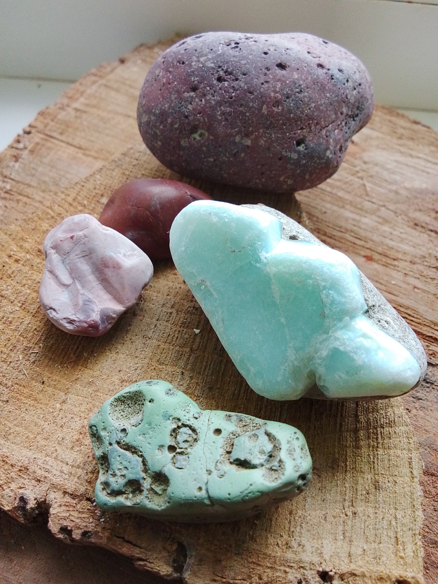 The image shows five stones of different sizes and colors arranged on a wooden surface. The surface appears to be a rough-cut piece of wood. The largest stone is dark reddish-purple with small holes. There is a smaller red stone next to a pink stone. A light blue-green stone is positioned prominently in the middle. The fifth stone, at the bottom, is a textured, green-greyish color with numerous small indentations. The image was likely taken in natural lighting. The location of 1-Go Maya, Russia is not apparent in the image.