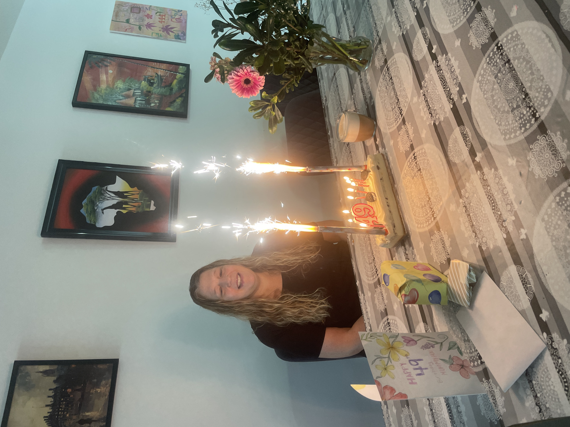 The image shows an indoor birthday celebration scene located in Raanana, Israel. A woman with long, wavy, light brown hair, wearing a dark short-sleeved top, is seated at a table, smiling. In front of her, on the table, is a birthday cake adorned with candles forming the number "49" and several smaller lit candles, flanked by two actively sparking celebratory candles emitting bright white sparks. To the left of the cake are a birthday card with floral designs, displaying "HAPPY 49th Birthday MUMMY!!", a yellow gift wrapped with multicolored balloon patterns, a smaller striped gift, and a white envelope. To the right of the cake, a glass vase holds a bouquet featuring a pink gerbera daisy and green foliage, alongside a glass containing a layered beverage, possibly a latte. The table is covered with a grey and white patterned tablecloth. The background consists of a plain light-colored wall displaying four framed art pieces: a dark, impressionistic city street scene, a vibrant depiction of giraffes and an acacia tree within an outline of the African continent, a framed painting of an exotic village with huts and a boat on water, and an abstract artwork with floral elements and numbers including "444".