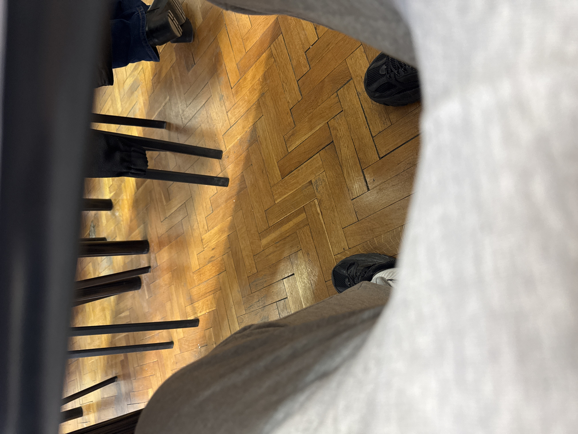 A first-person perspective photograph taken indoors showing a parquet wood floor, multiple chairs, and portions of two individuals. One person is wearing black shoes and gray sweatpants. Another person is wearing a black dress, blue jeans, and black leather shoes. The location is likely in a building in Sofia, Bulgaria, based on the presence of parquet flooring, a common feature in Bulgarian architecture. The floor has a herringbone pattern.