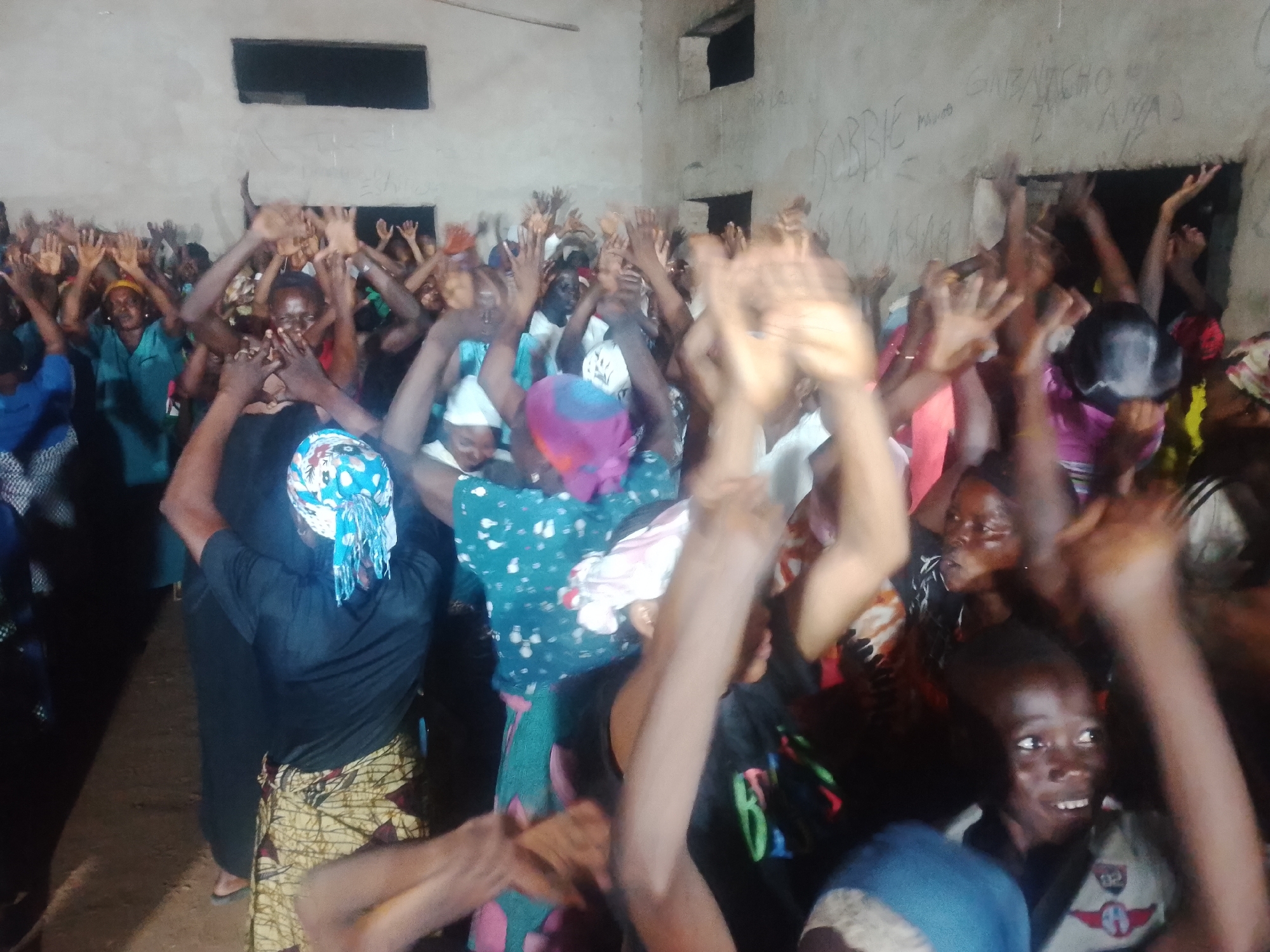 In an indoor setting with a concrete floor and walls, a group of individuals, predominantly women and children, are raising their hands in the air. The room appears to be a community hall or a similar gathering space, with a dark rectangular opening in the wall, possibly a window or vent, and another similar opening in the far wall. Graffiti is visible on the wall in the background. The individuals are dressed in casual clothing, with some wearing headscarves. The atmosphere suggests a shared emotional experience, possibly a religious service or a community event.