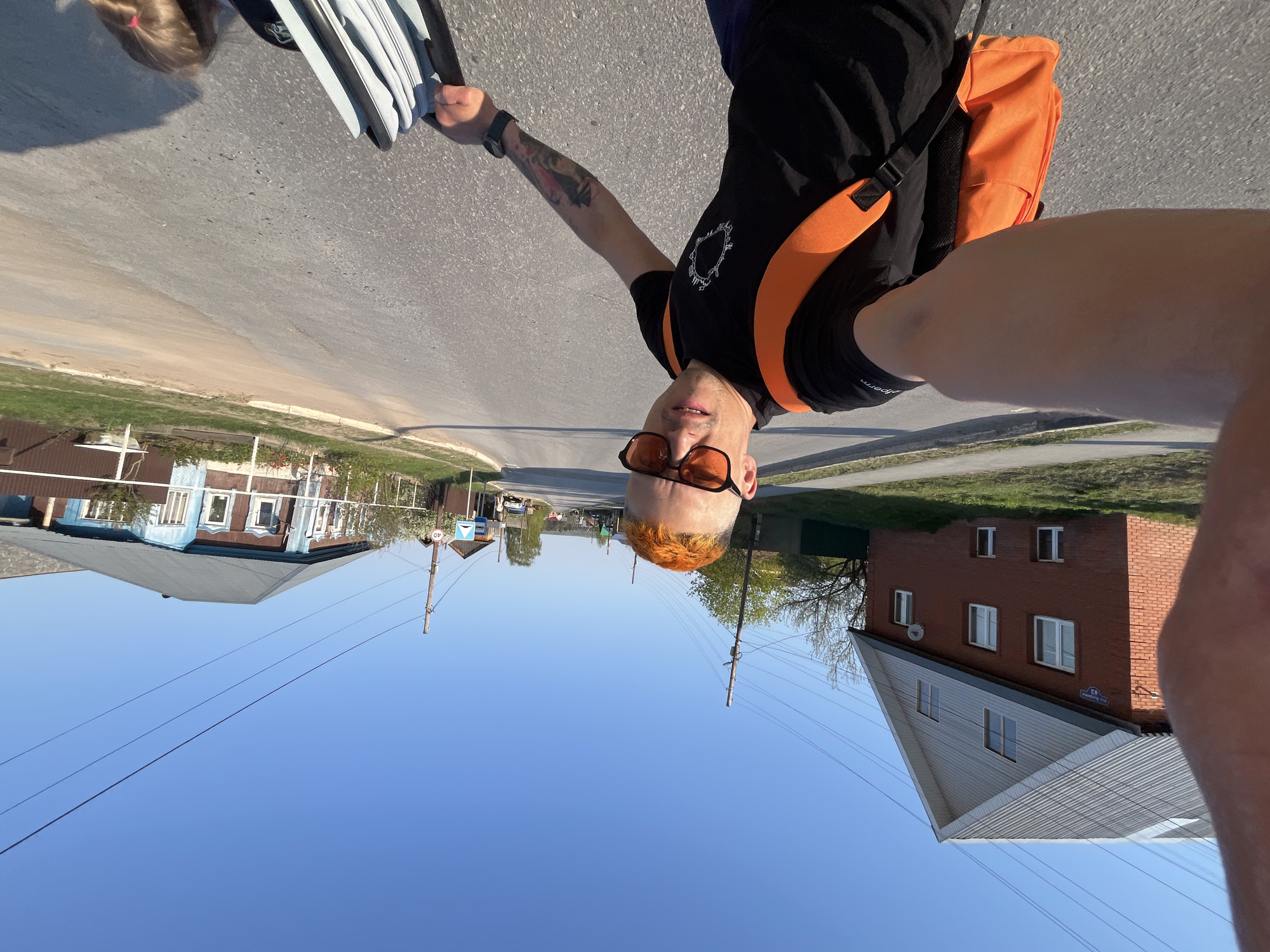 A selfie taken in the city of Chaikovsky, Russia, shows a fair-skinned person with orange hair and sunglasses, wearing a black t-shirt with an orange backpack. The person is holding what appears to be a white car seat cover. The background depicts a typical Russian street with houses and power lines. A road sign indicating a speed limit of 40 is visible. A portion of another person's head is visible at the top left. The building at the right has a street sign with the number 42.