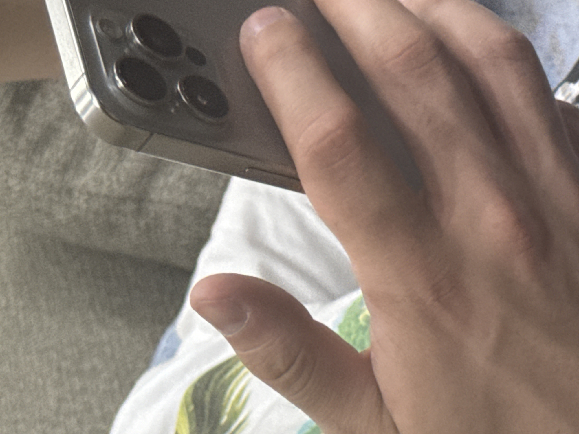 The image shows a close-up view of a person's hands holding a silver smartphone. The phone has a triple-camera system on the back. The person is wearing a white t-shirt with a pattern of green leaves and blue elements. The background is blurred, showing a textured grey surface, likely a piece of furniture or clothing. There are no identifiable landmarks or context to suggest the location is Tbilisi, Georgia. The image focuses on the hands and the phone, with the background providing minimal environmental context.