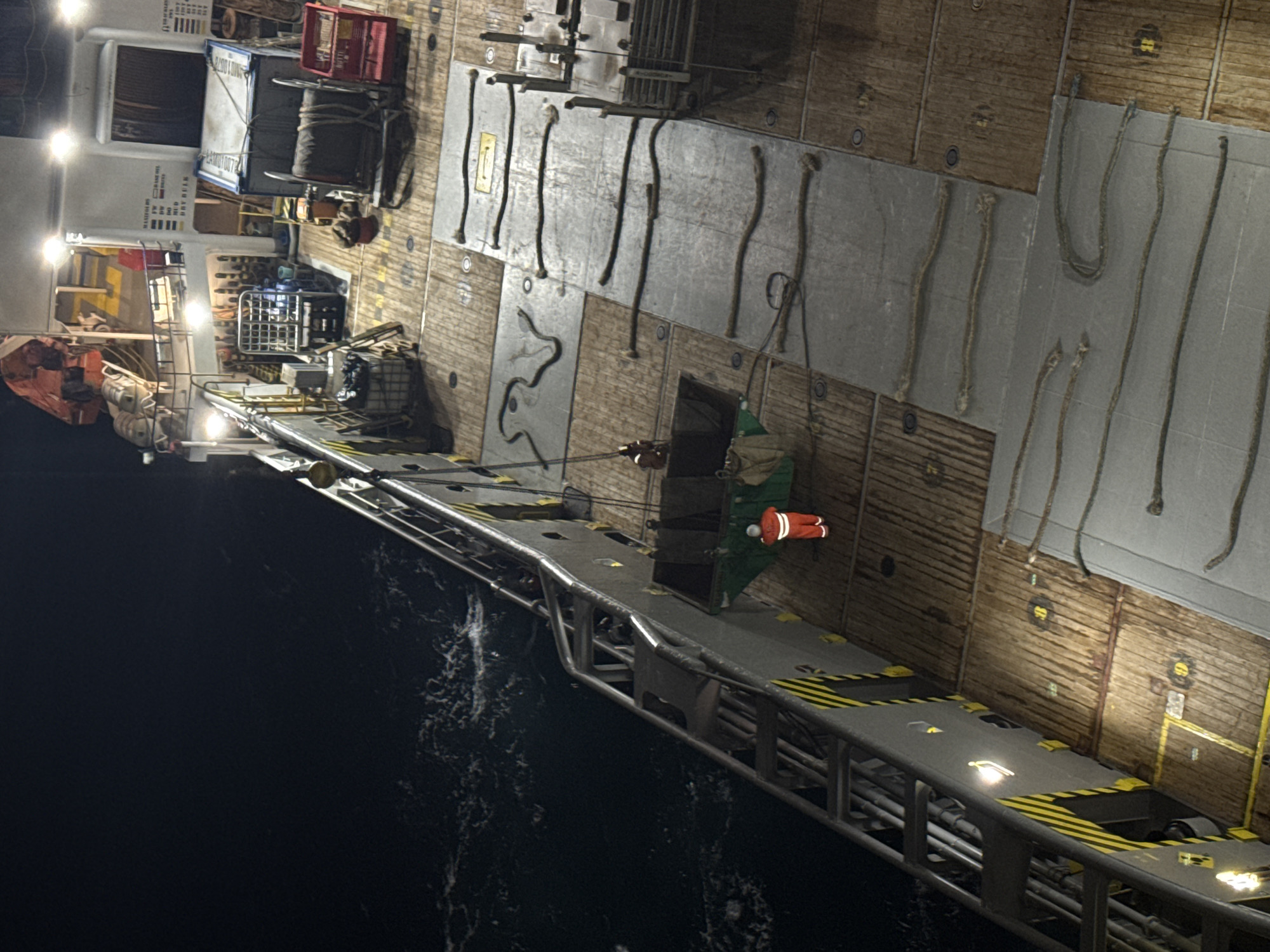 The image depicts the deck of a ship at night, with a person in an orange jumpsuit standing near a large green bin. The deck is made of wood and metal plating, and various ropes are coiled and laid out. In the background, there are containers, machinery, and lights illuminating the scene. The water is visible to the left of the ship, with some waves. There are no identifiable landmarks or clues to suggest a specific location within a city, let alone Unknown, Italy.