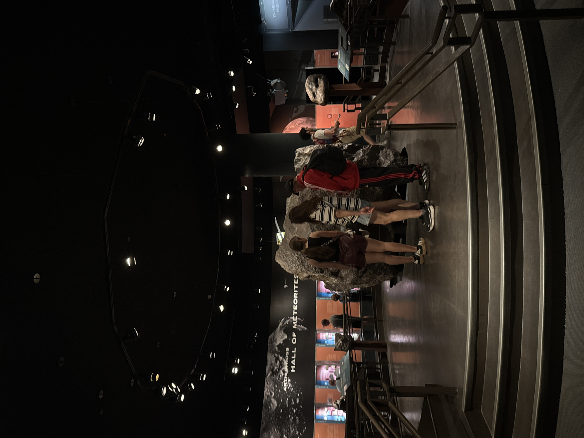 This image captures an indoor scene within a museum exhibition hall, specifically identified as the "Arthur Ross Hall of Meteorites." The overall setting is dimly lit with targeted spotlights illuminating the exhibits, creating a dramatic and immersive atmosphere.
In the mid-ground, several people are gathered around and observing large, irregular rock formations, which are prominently displayed meteorites. One person, seen from behind, wears a red jacket, black track pants with white stripes, and a baseball cap. Another individual wears a striped top and shorts, while a third is in a dark top and shorts. They appear engaged with the exhibits, suggesting an educational visit.
To the right of the frame, a wide, tiered set of steps or seating area with a metal handrail ascends. Along the far wall, large display screens and information panels are visible, featuring images and text. The most prominent visible text is a large sign that reads "ARTHUR ROSS HALL OF METEORITES." Below this, smaller, less legible signs on display cases appear to reference geological or cosmic layers, with words like "CRUST," "MANTLE," and "CORE" faintly discernible. A model of a celestial body or spacecraft can be seen suspended from the high, dark ceiling in the background.
The scene depicts museum visitors actively exploring and learning within a specialized exhibit, likely a natural history or science museum in New York, United States of America. The time of day is indeterminate due to the artificial lighting.