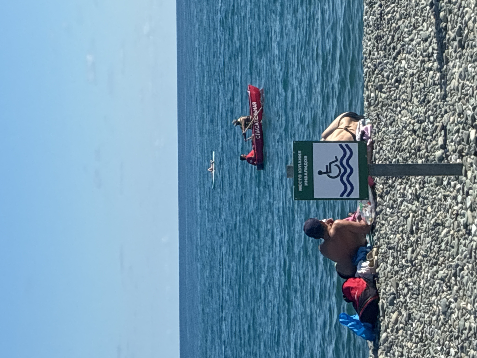 A wide shot captures a sunny day at a pebble beach in Sirius, Russia, bordering the Black Sea. The sea is a vibrant turquoise, with gentle waves under a clear blue sky. In the foreground, two individuals are relaxing on the shore. One person, facing away from the camera, wears a dark cap and appears to be lying on their back, while the other, a woman wearing a bikini, is partially visible in profile. Beach gear, including a blue plastic bag, a red backpack, and a blanket, is scattered near them. A sign with a wheelchair symbol and Russian text indicating a "place for swimming for people with disabilities" stands prominently on the beach. In the middle ground of the sea, a small red rescue boat with two occupants is visible, one rowing. Further out, a person on a paddleboard is also present. The scene conveys a sense of leisure and the presence of safety measures for beachgoers.