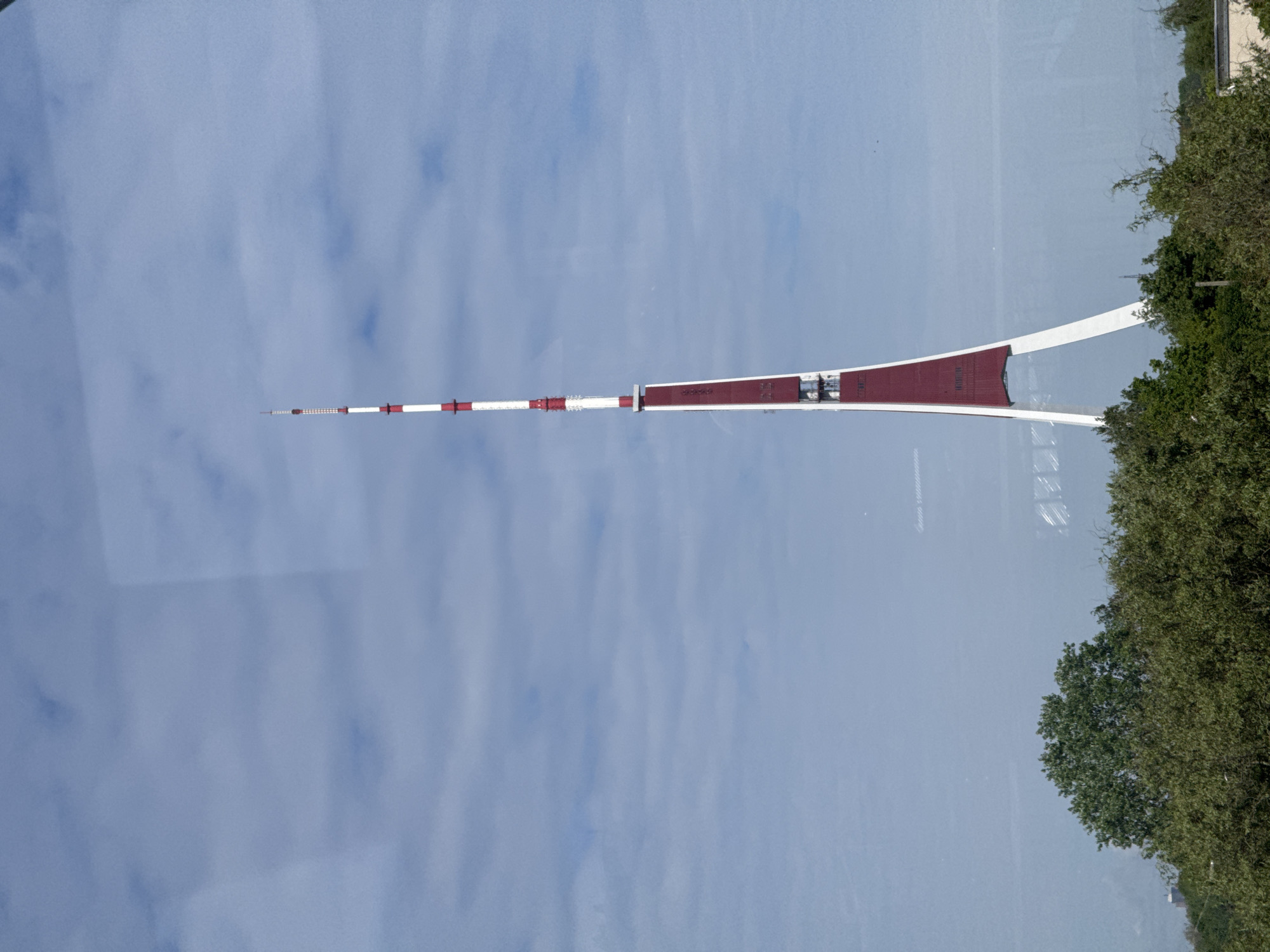 The image depicts the Riga Radio and TV Tower, a prominent landmark in Riga, Latvia, seen from an elevated perspective. The tower features a slender spire alternating with red and white horizontal bands. The base is broader with a reddish-brown color. The background is a cloudy sky. Part of the tower structure appears to reflect in glass in the immediate foreground, with trees visible on the right side of the frame.