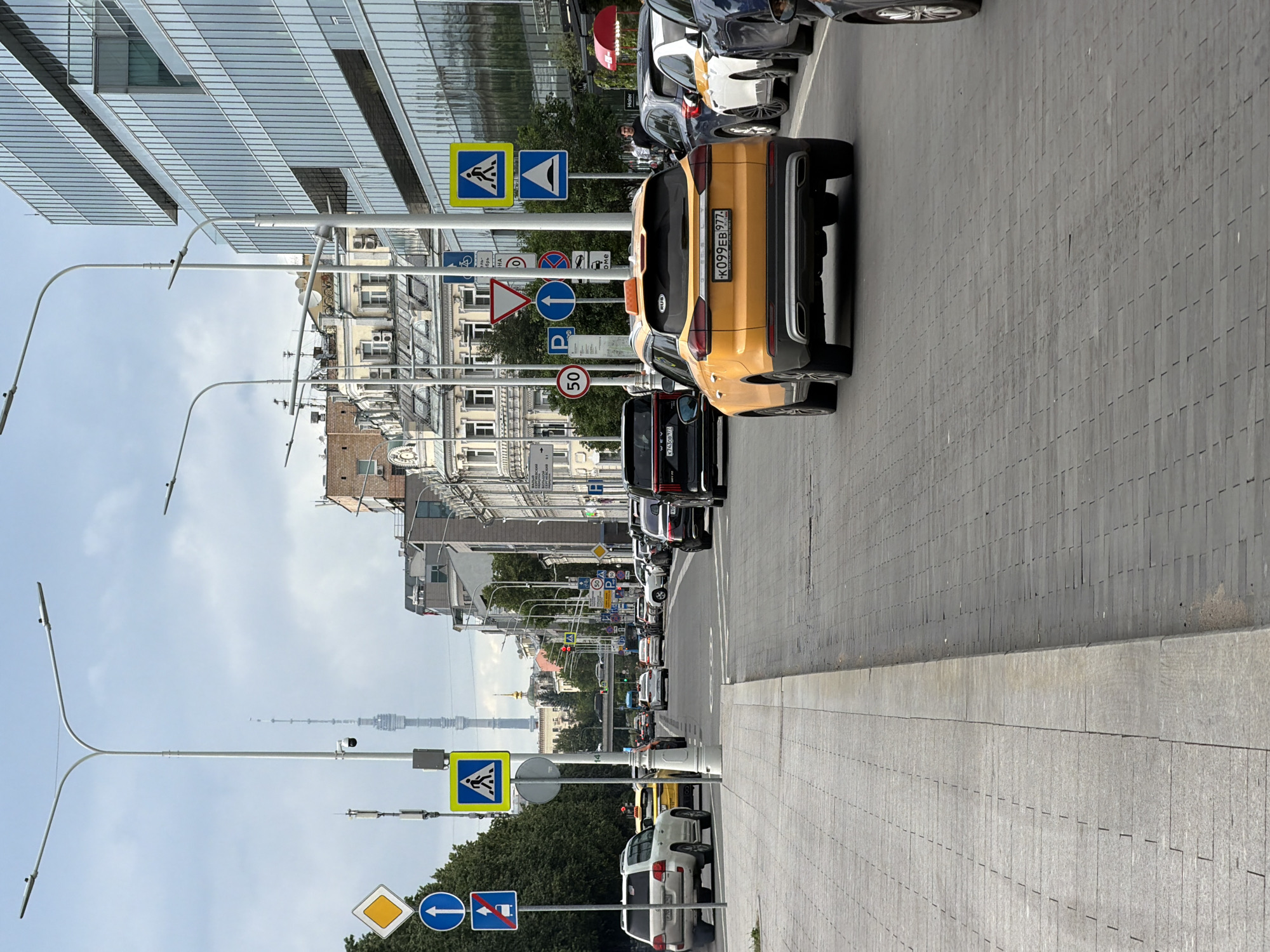 The image depicts a street scene in an urban environment, with a focus on traffic and pedestrian infrastructure. Several cars are parked and in motion on the right side of the street, with a prominent yellow car in the foreground. Traffic signs, including pedestrian crossings, speed limits (50), and directional arrows, are visible along the sidewalk. A modern glass-fronted building dominates the left side of the frame, contrasting with older, multi-story buildings further down the street. Streetlights with a modern design are also present. In the distance, a tall, lattice-like tower is visible against a cloudy sky, suggesting a specific landmark. The scene appears to be during daylight, with overcast weather conditions. The overall impression is one of everyday urban activity, with clear signage and a mix of architectural styles. No people are clearly identifiable in the image. Visible text includes license plates, such as "K099EB 977" and "K743XB 777", and markings on some traffic signs.