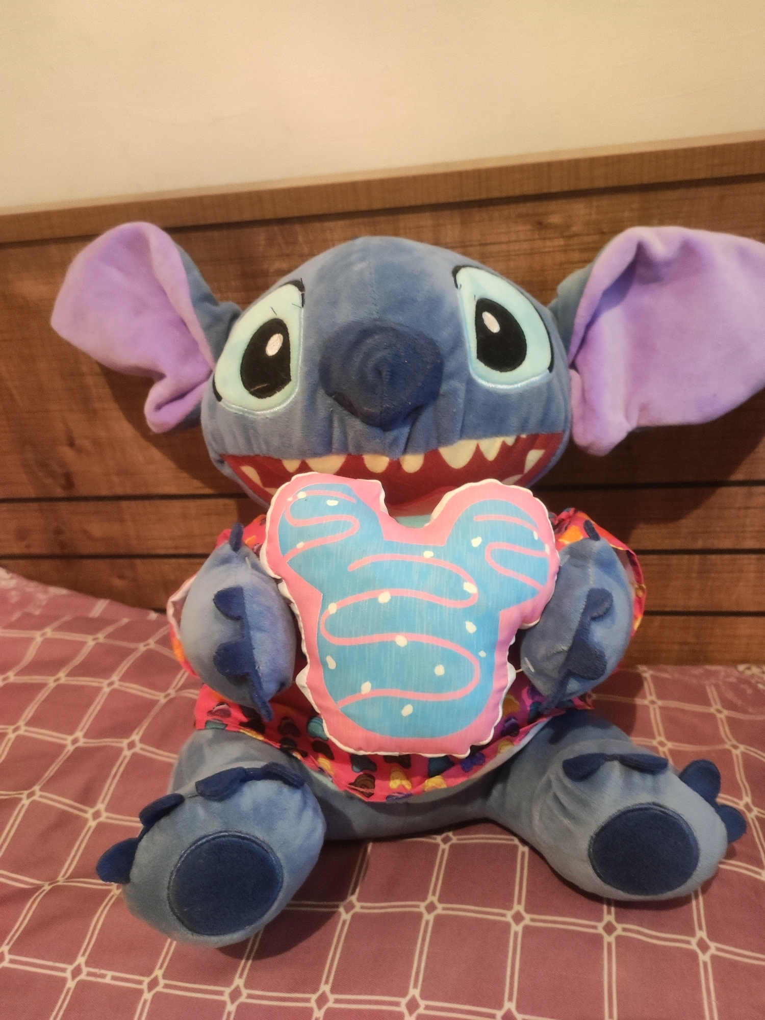 A plush toy Stitch character is sitting on a bed. The toy is blue with large purple ears and a pink and blue Mickey Mouse-shaped pillow. The bed has a purple and white checkered blanket. The background appears to be a wooden wall. The image is taken at an eye-level perspective. The lighting is consistent throughout the scene. There is no indication of the location being Khazar Raion, Azerbaijan.