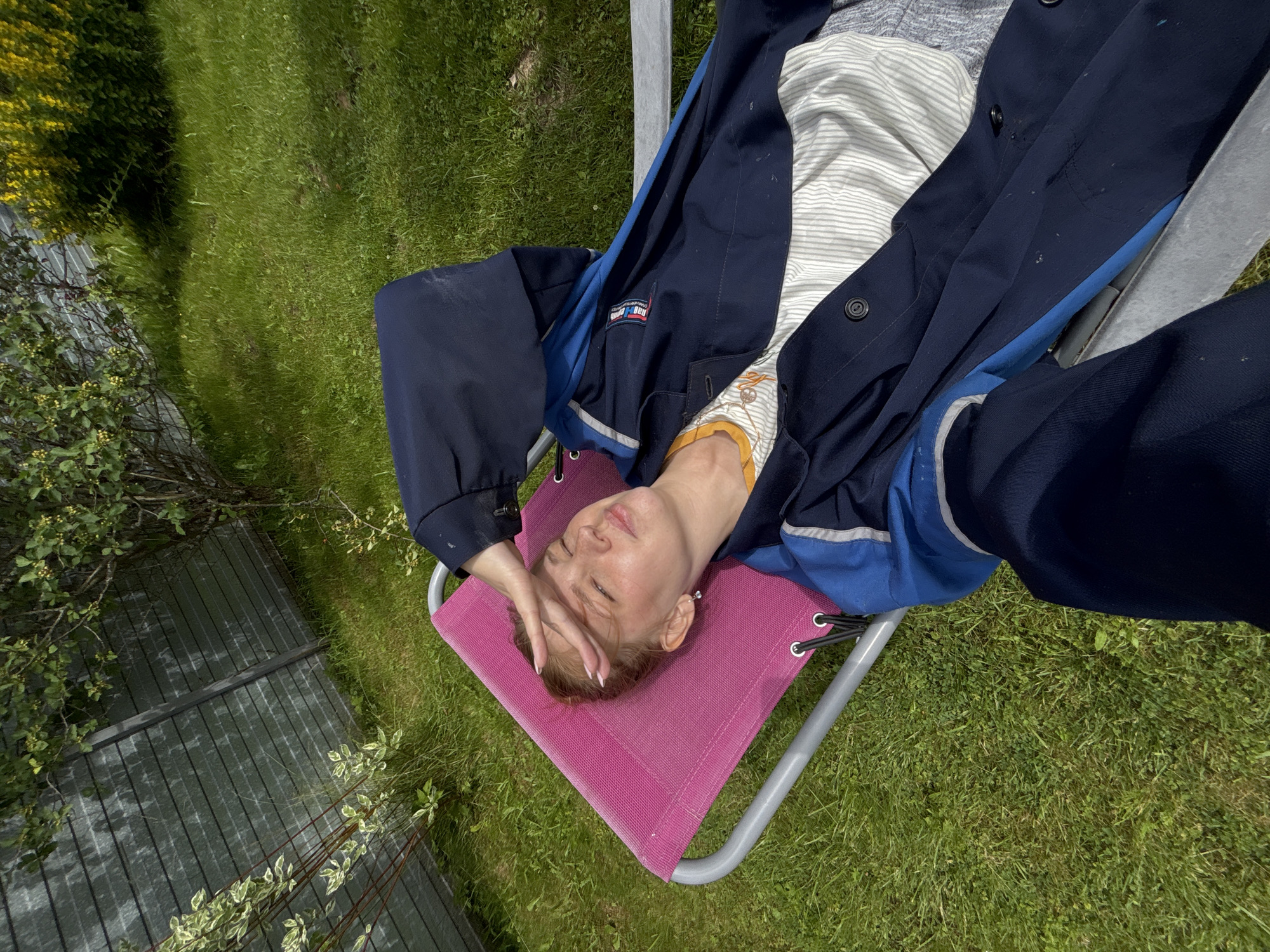The image shows a young woman lying on a pink chaise lounge in a grassy backyard. She is wearing a dark blue jacket over a striped shirt and has her hand over her forehead, shielding her eyes from the sun. The background features a wooden fence and trees. The location is identified as Dunilovo, Russia.