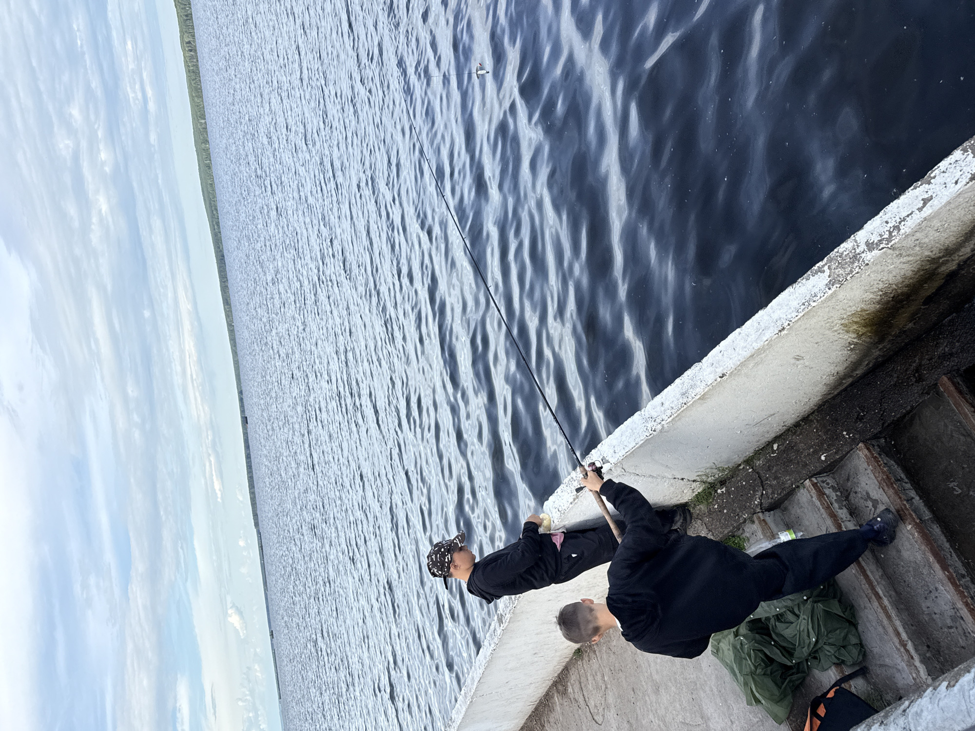 Two young individuals are fishing from a concrete embankment along a large body of water. The person closer to the viewer, facing away, is wearing a black hoodie and dark pants. The second individual, further away and facing the water, is wearing a black hoodie and a patterned cap. Both are holding fishing rods. The water is dark blue with ripples, and the sky is overcast with scattered clouds. The embankment is weathered and appears to be part of a man-made structure. To the left of the embankment, green bags and orange objects are visible near a set of concrete stairs leading down. The horizon in the distance shows a treeline. This scene likely depicts a leisure activity at a reservoir or large lake within Chaikovsky, Russia.