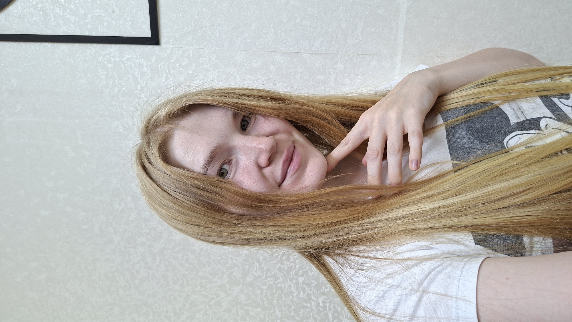 A young woman with long, blonde hair is smiling and looking at the camera. She is wearing a white t-shirt with a graphic print. Her right hand is resting on her chin. The background is a textured, light-colored wall. There is a framed picture on the wall to the right of the woman. The scene appears to be indoors, in a residential setting, potentially in a bedroom or living area. The location in Bolshoy Bukor, Russia cannot be determined from the image.