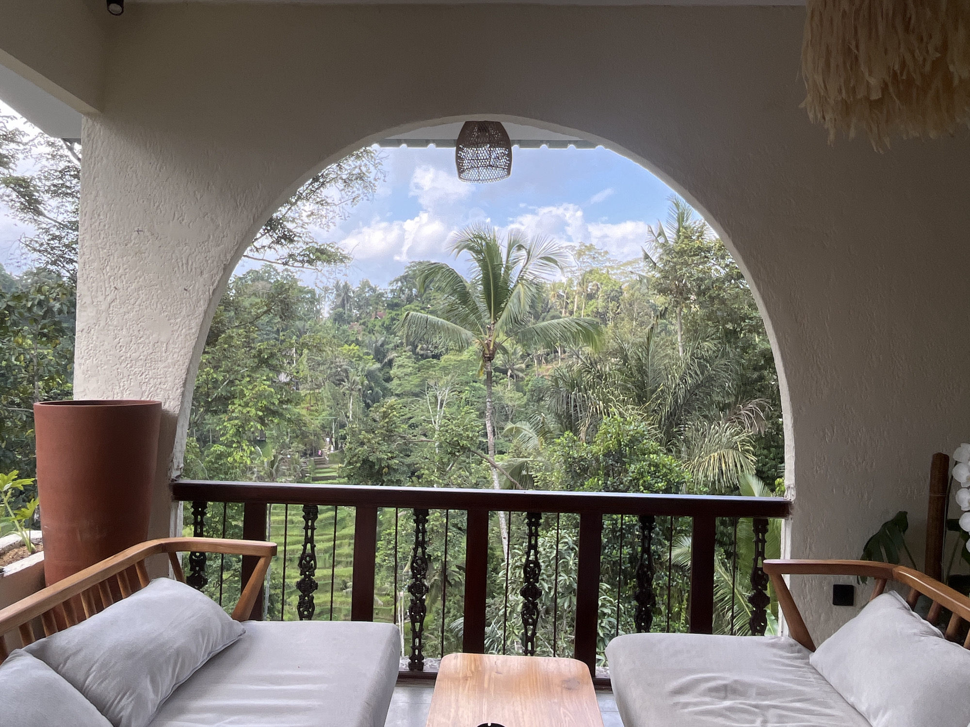 The image presents a tranquil scene from a balcony or veranda in Tegallalang, Indonesia, overlooking a lush tropical landscape. The view is framed by a wide, textured, light-colored arched opening.
In the foreground, two comfortable daybeds with light grey cushions and pillows are arranged facing outward, separated by a small, light-colored wooden table. A dark wooden railing with ornate metal balusters runs across the width of the balcony. On the left, a tall, terracotta-colored pot stands next to the balustrade, while on the right, hints of white flowers (possibly orchids) are visible near the right daybed. The walls and arch have a rough, stucco-like texture. A small, black light fixture is visible on the ceiling of the archway on the left.
Through the arched opening, a vibrant green valley of dense tropical foliage dominates the view. Numerous palm trees, including a prominent one in the center, rise above the canopy. Beyond the immediate jungle, glimpses of terraced rice paddies can be seen in the distance, characteristic of the Tegallalang area. The sky above is bright blue with scattered white clouds, indicating daytime and pleasant weather conditions.
A woven, basket-like lantern hangs from the center of the arched ceiling, and a light-colored, fringed decorative fixture is suspended from the ceiling on the right side. The scene conveys a sense of peaceful relaxation and a close connection to nature. There are no people visible, and no visible text in the image.