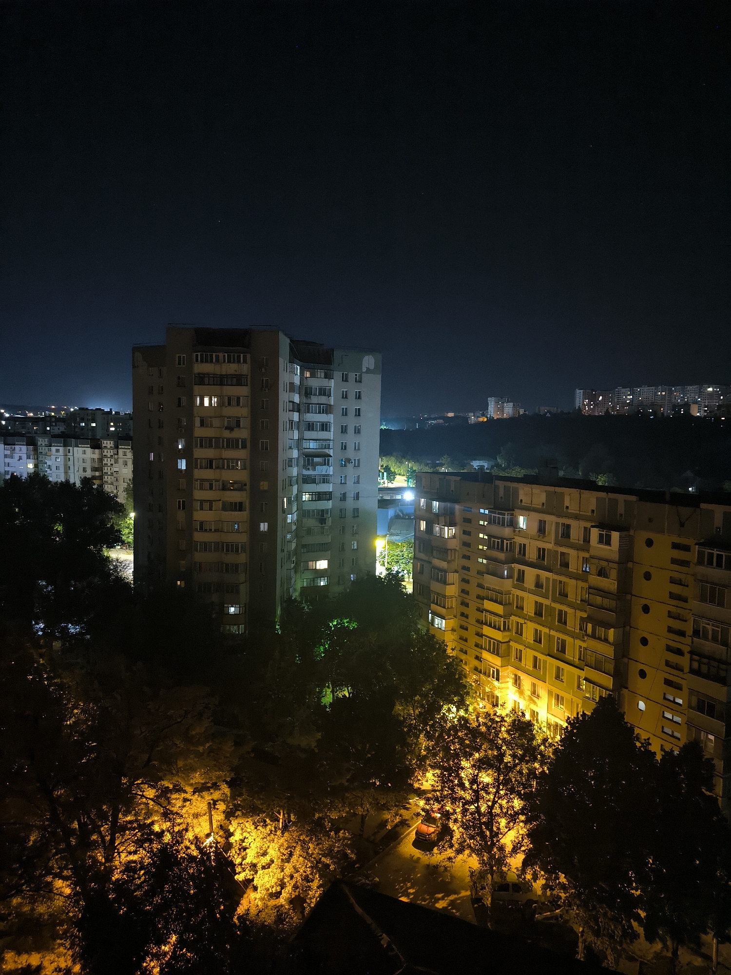 The image depicts a nighttime aerial view of a residential area in Chișinău, Moldova. Dominating the foreground are several multi-story apartment buildings, illuminated by the warm glow of interior lights and street lamps. Trees, some with foliage brightly lit by the artificial illumination, are interspersed between the buildings and in the surrounding areas, creating a stark contrast between light and shadow. In the background, more apartment buildings and city lights extend into the distance, suggesting a densely populated urban environment. The sky is dark, with a few faint stars visible. The scene captures the typical appearance of a city at night, with a focus on the architecture and natural elements illuminated by human-made light sources. There are no people or vehicles clearly visible or identifiable.
