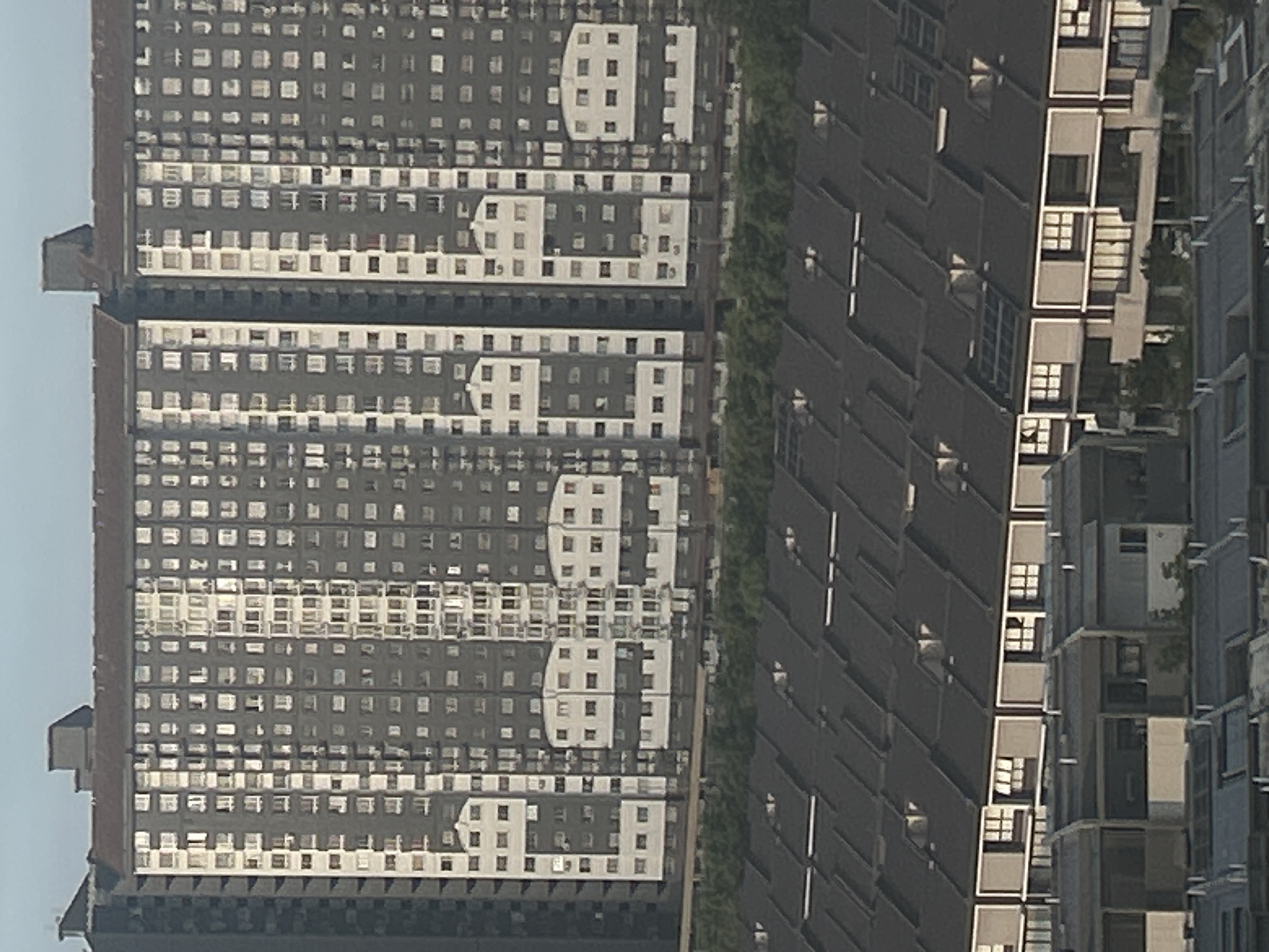 The image depicts a dense urban scene, likely a residential area in Kuala Lumpur, Malaysia, given the architectural style of the buildings. Two high-rise apartment buildings dominate the view. The building on the left has a grid-like facade with numerous windows, arranged in a repetitive pattern. The color scheme is predominantly gray and white. The building on the right consists of angular, darker-colored modules, each featuring a balcony or open space. A line of dense greenery separates the two buildings, implying a street or park area. The photograph appears to be taken during daylight hours, judging by the brightness. There are no people visible in the image.