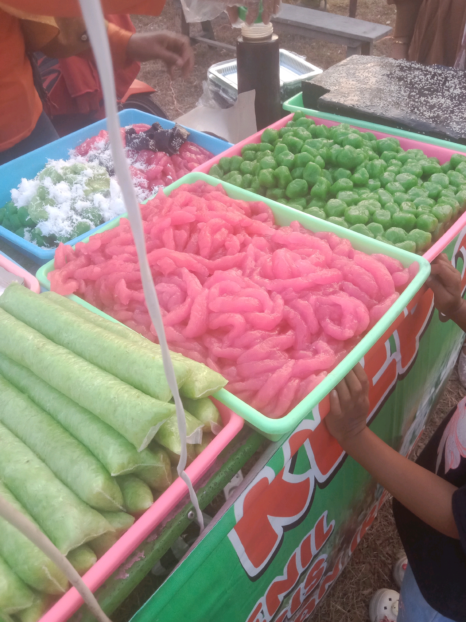 The image shows a street food stall in Johore, Indonesia, displaying a variety of colorful desserts. In the foreground, a person's arm and hand reach out to the stall. The stall itself is colorful, with green, pink, and orange accents.
The food items on display include:
* Green, cylindrical items in plastic packaging, arranged in rows.
* Pink, ribbon-like dessert in a green plastic tray.
* Small, round green candies in a pink plastic tray.
* A blue tray with a mix of green and white shredded coconut-like dessert, and some dark, granular toppings.
* A dark, rectangular cake-like item covered in sesame seeds, sitting in a green tray.
* A dark bottle with a dispenser cap, possibly for syrup or sauce.
The background shows a wooden bench and a person in an orange shirt. The ground appears to be dirt and dry grass. The overall impression is a bustling street food scene.