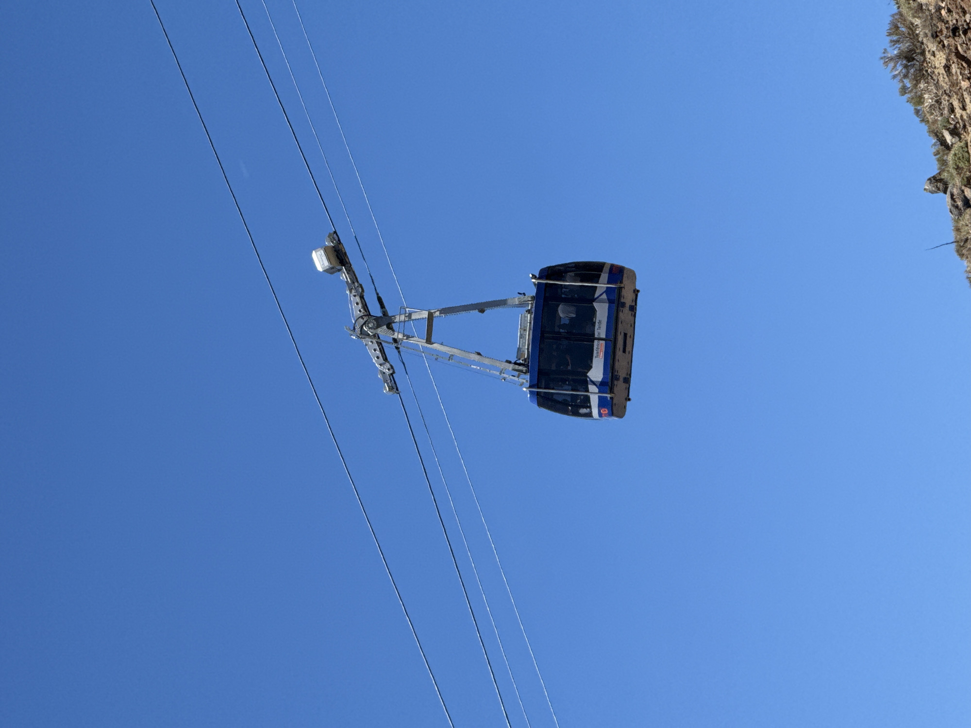 A blue and white cable car is suspended in the air against a clear blue sky. It is attached to a metallic support structure that runs along several thick wires, which extend across the frame. The cable car has tinted windows and appears to be in motion. The bottom portion of the image shows a rocky, arid landscape with sparse vegetation. The cable car appears to be part of the Teleférico del Teide, a cable car system that ascends to the summit of Mount Teide, located in the Canary Islands, Spain, which is accessible from the town of La Orotava.