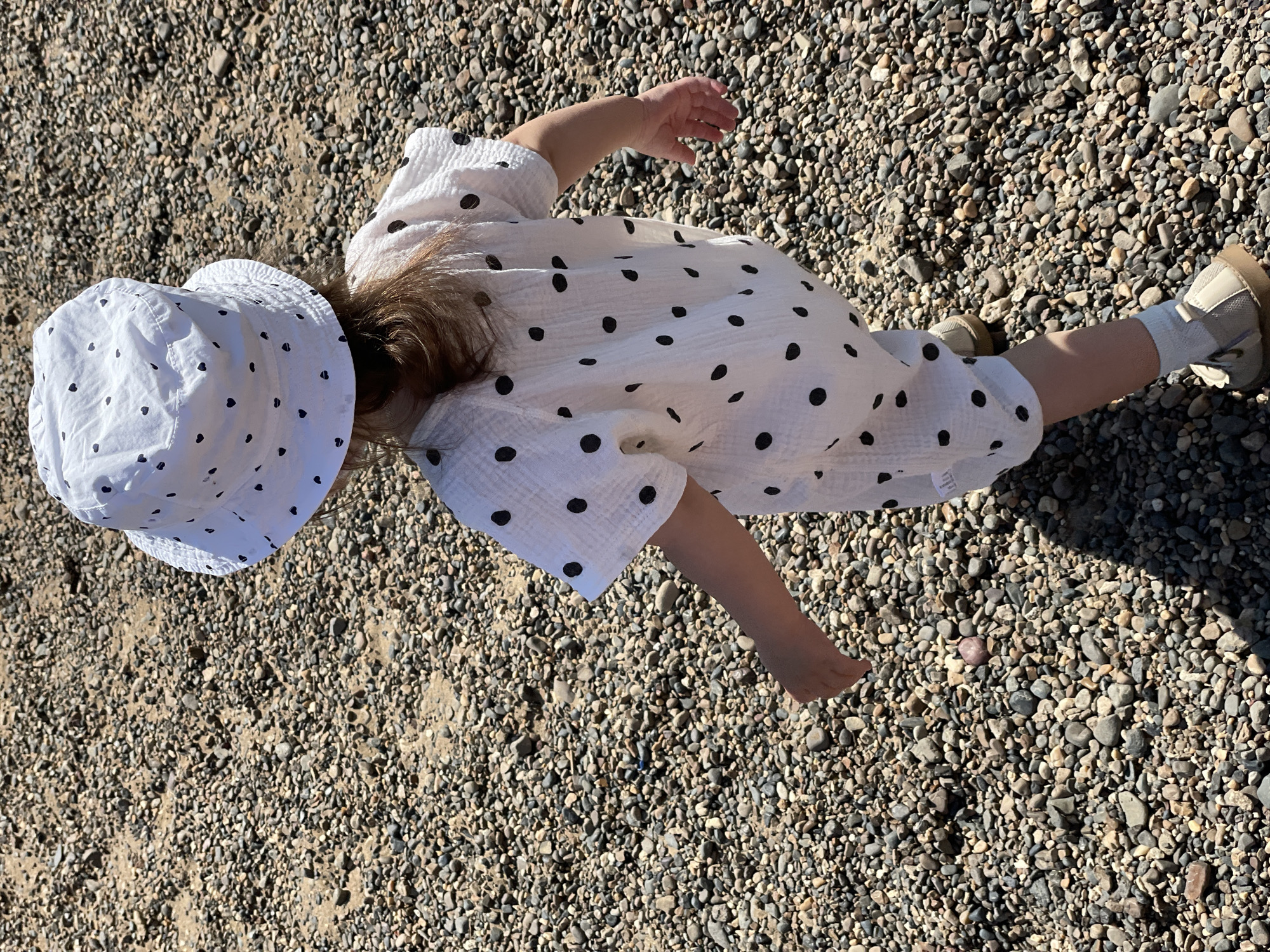 A child wearing a white polka-dot romper and matching bucket hat lies on a pebble-covered surface. The child is wearing socks and shoes. The location is unknown, and there is no information to suggest it is in Izhevsk, Russia.