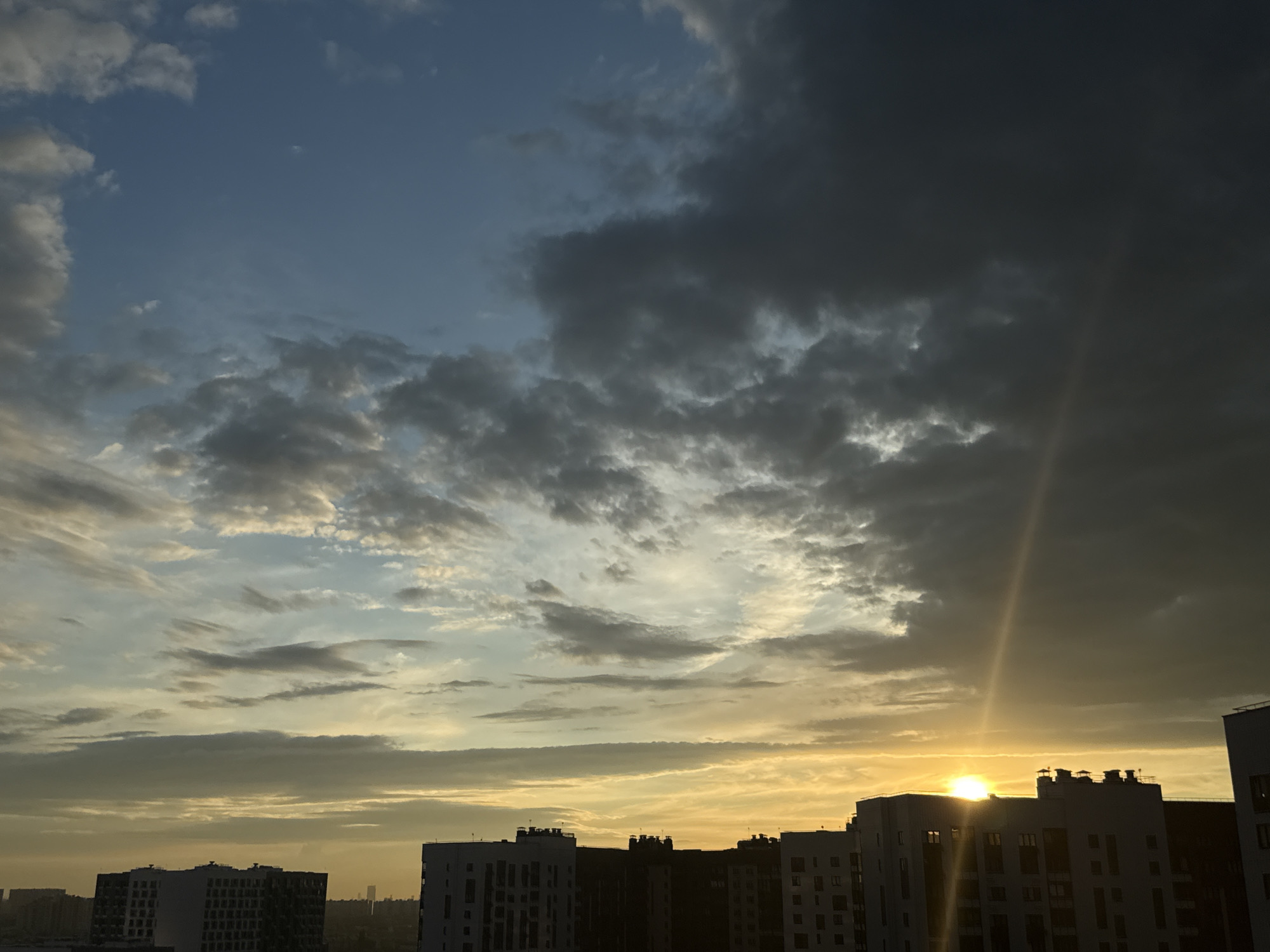 The image depicts a cityscape at sunset in Lyubertsy, Russia. A dramatic sky dominates the scene, filled with a mix of dark, heavy clouds on the right and lighter, more wispy clouds on the left, illuminated by the setting sun. The sun is a bright, glowing orb low on the horizon, casting a strong sunbeam that streaks diagonally across the darker clouds. Silhouetted against the colorful sky are several tall, modern apartment buildings, characteristic of urban residential architecture. The buildings are predominantly light-colored, with visible windows and rooftop details. In the distance, the urban landscape extends, with hints of other structures. The overall impression is one of an urban environment transitioning into evening, with the sky being the most striking element.