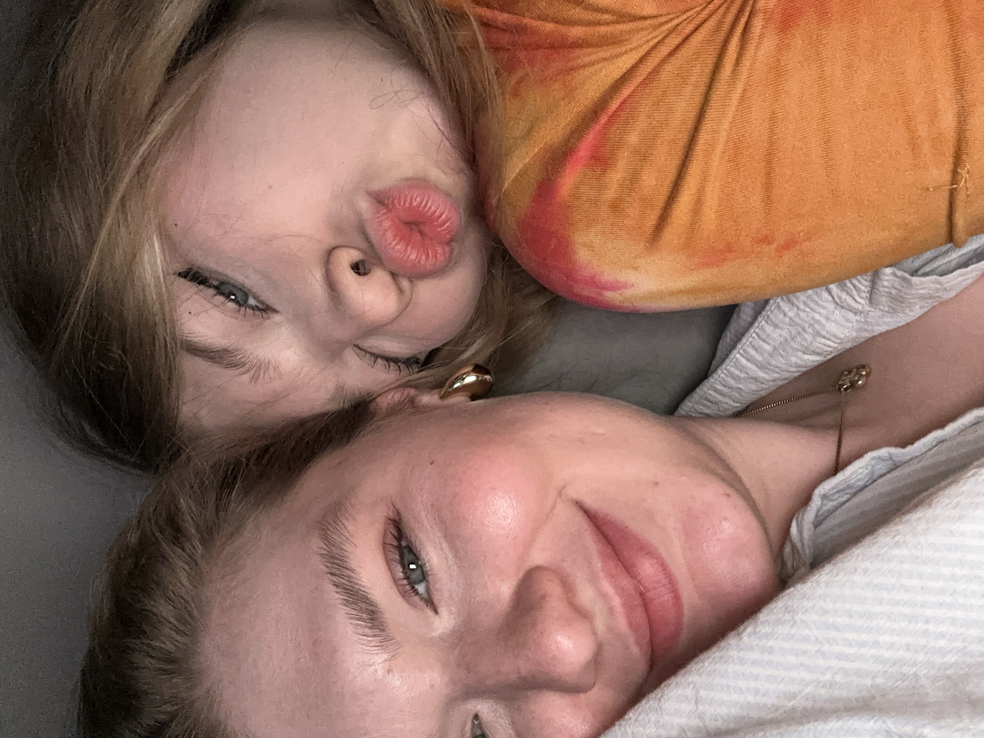 Two people are lying down next to each other. The person on the top is pouting their lips. The person on the bottom is smiling slightly. There is an orange tie-dye shirt visible. A small gold chain with a pendant is visible on the person on the bottom. The location is not specified.