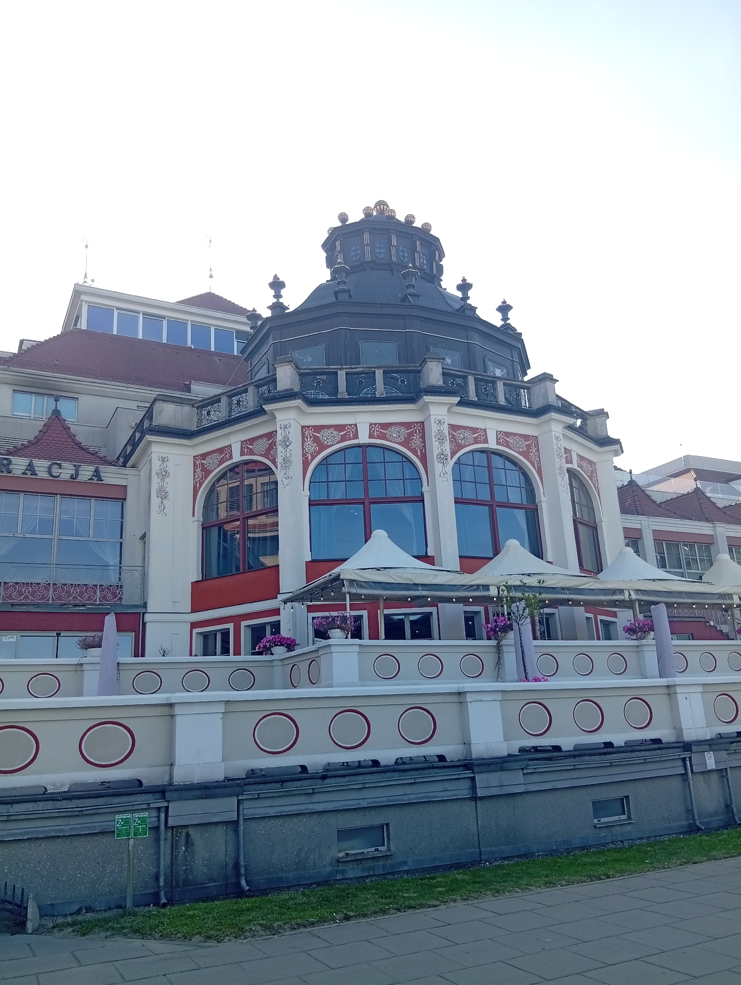 The image shows the historic Sopot Pier in Sopot, Poland, on a clear day with a bright sky. The iconic building at the pier entrance, the Grand Hotel, is prominently featured. The building exhibits a distinctive architectural style with a large, ornate dome topped with a crown-like structure. The facade is painted in white and red, with large arched windows framed by decorative white detailing. Several white umbrellas are set up on a terrace in front of the building, suggesting an outdoor seating area for a cafe or restaurant. Potted plants with pink flowers are placed along the railing of the terrace. The lower part of the building has a cream-colored wall with a repeating pattern of red-rimmed circles. Below the wall, a concrete structure with small windows and drainage pipes is visible, leading down to a paved walkway and a strip of grass. A small green sign with white text and icons is fixed to the concrete wall. No people are visible in the image.