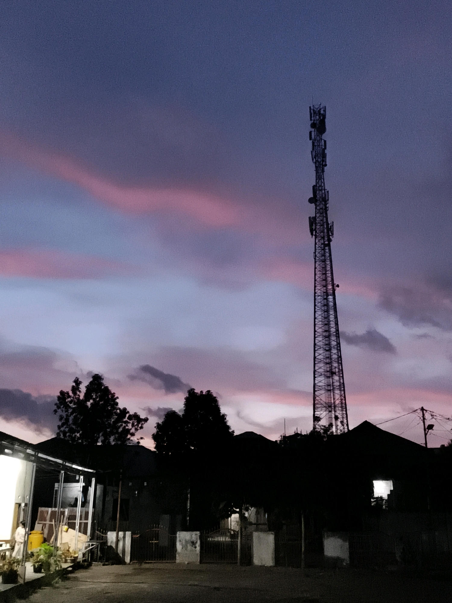 At dusk in Tombatu Dua, Indonesia, a tall, lattice-style telecommunications tower dominates the skyline, silhouetted against a sky streaked with pale pink and lavender clouds. Below the tower, the dark shapes of trees and residential buildings are visible. A single light shines from a window in one of the houses. In the foreground, a paved area shows faint outlines of parked objects and a person standing near a yellow barrel and some potted plants. The scene captures a quiet moment as daylight fades.