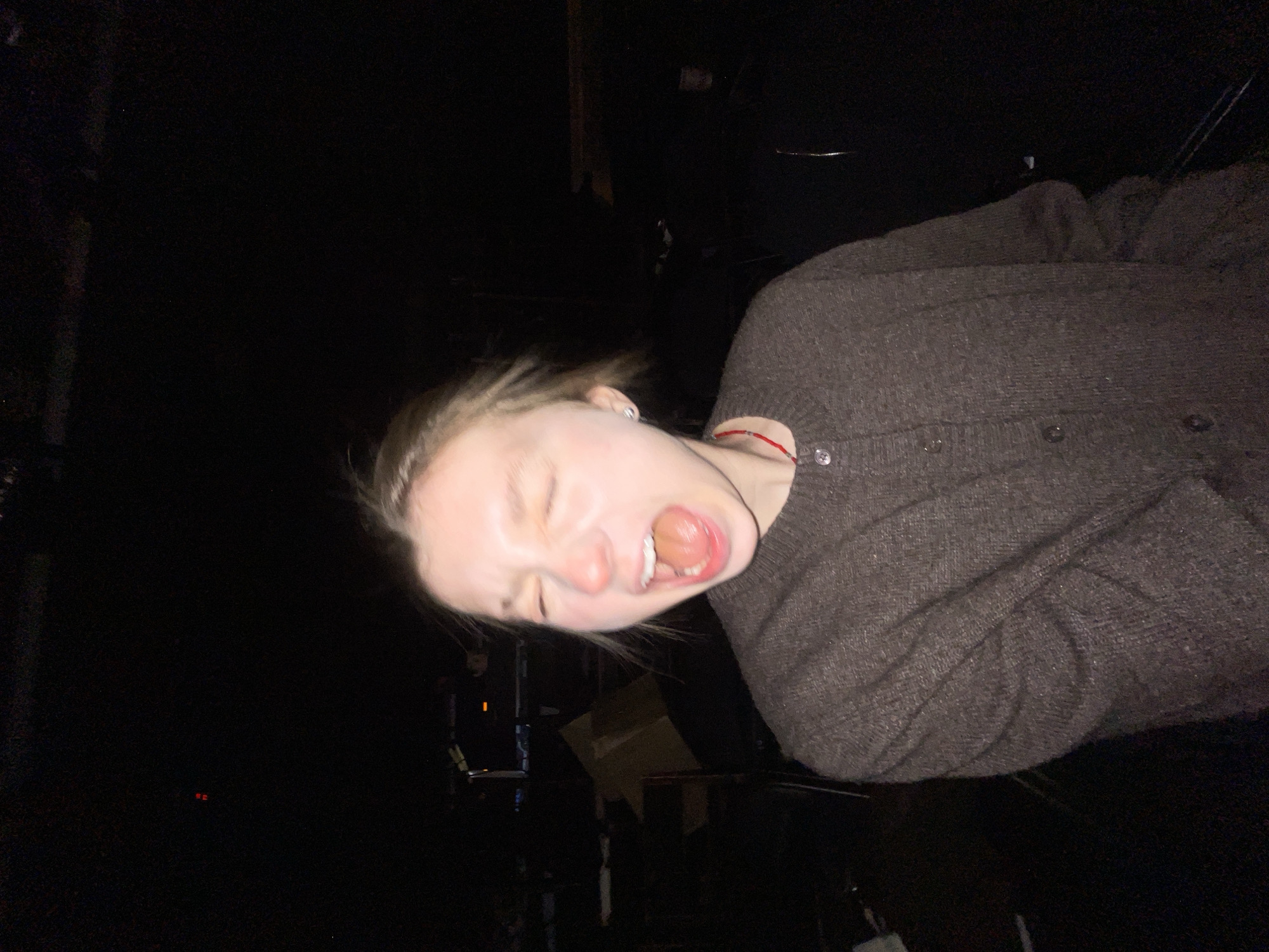 A young, light-skinned person is seen in a tilted, blurry photo taken at night. The individual has fair hair, is wearing a dark-colored cardigan, and is screaming with their mouth wide open. In the background, indistinct shapes and shadows suggest an urban environment at night, potentially in Moscow, Russia given the prompt. There is a hint of structural elements, possibly buildings or urban features, in the dark background, and what may be a cardboard box. The image is poorly lit and taken from a low angle, emphasizing the person's face and upper body.