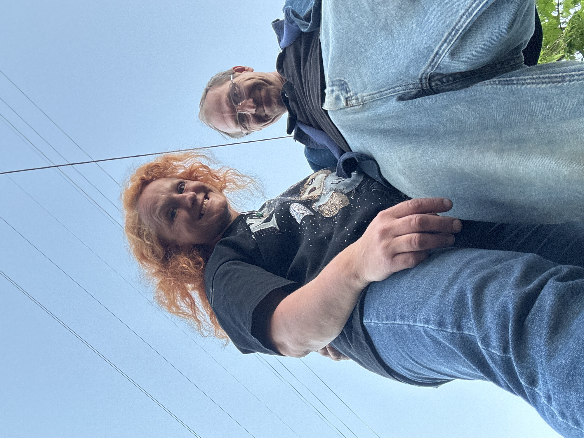 An unusual low-angle photograph captures two smiling adults looking down at the camera against a clear, bright blue sky, likely taken in Chaikovsky, Russia. The perspective is from directly below the subjects, giving a worm's-eye view.
On the left, an adult female with striking red, curly hair and a wide smile is visible. She wears a black t-shirt adorned with a shiny, possibly sequined or glittery, design that appears to feature an animal or abstract pattern. Her left arm is bent, and she wears blue jeans.
To her right, an adult male with graying hair and glasses is also smiling down. He is dressed in a dark shirt underneath a blue denim jacket and blue jeans. His body is partially obscured by the female and the unusual angle.
In the background, several parallel power lines cut across the clear blue sky. A hint of green foliage, likely from trees, is visible in the upper right corner, suggesting an outdoor setting. The overall mood conveyed by the subjects' smiles is cheerful and relaxed. There is no visible text in the image.
