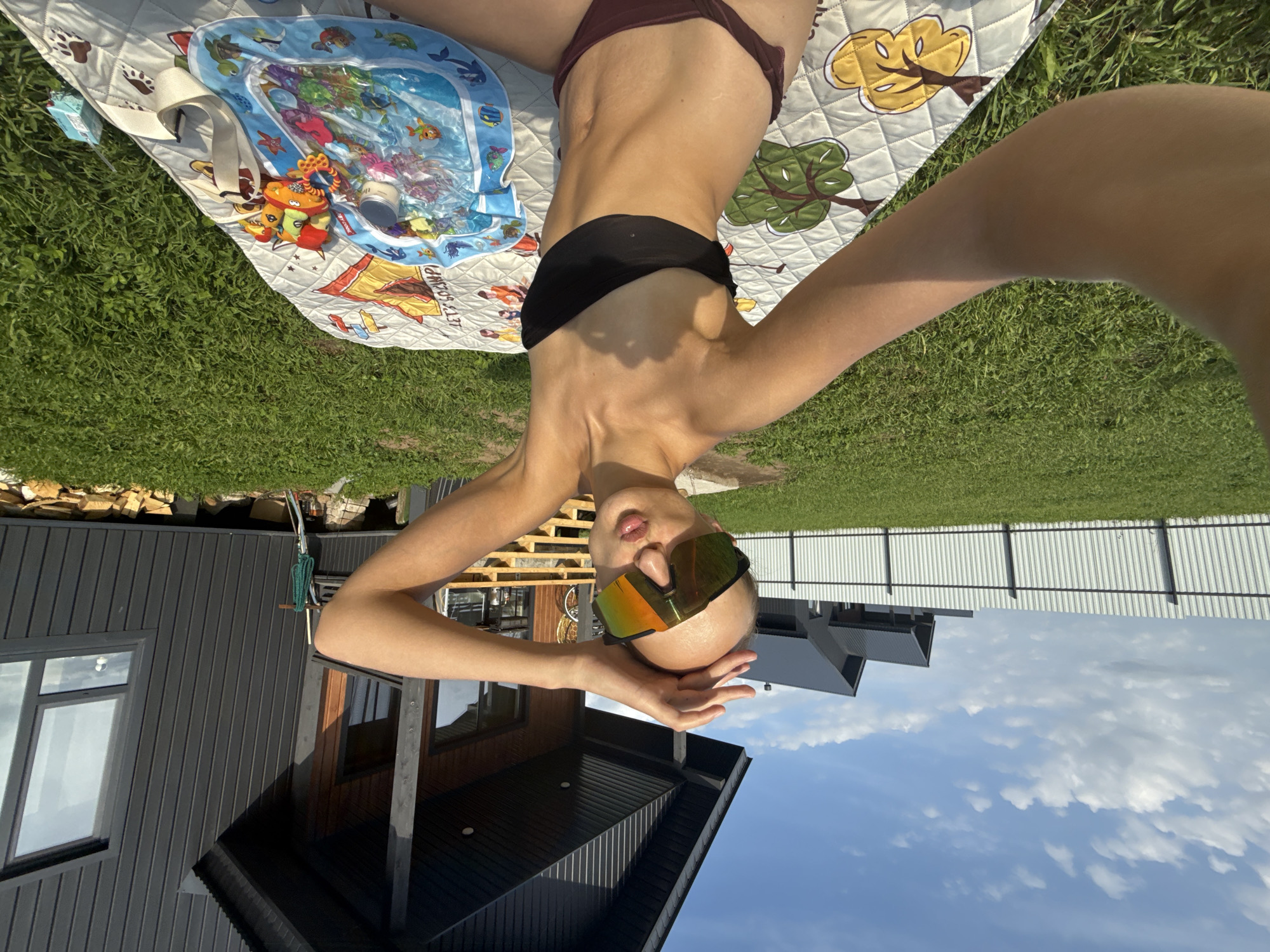 A young woman with a shaved head, wearing large orange-tinted ski goggles and a black strapless bikini top with maroon bottoms, is taking a selfie outdoors. She is sitting on a patterned picnic blanket on a green grassy lawn in front of a modern, dark-colored house with wooden accents and a porch. The house has large windows and a sloped roof. Behind the house, there is a white corrugated metal fence, and further in the distance, more houses with dark roofs are visible under a blue sky with scattered white clouds. On the blanket next to the woman is an inflatable water toy with sea creatures, a small jar, and other objects. The woman is raising one hand to her head, and her lips are pursed as if blowing a kiss. The scene suggests a sunny day in a suburban or rural setting, possibly the village of Podshivalovo, Russia, given the architectural style and the presence of a dacha-like house.