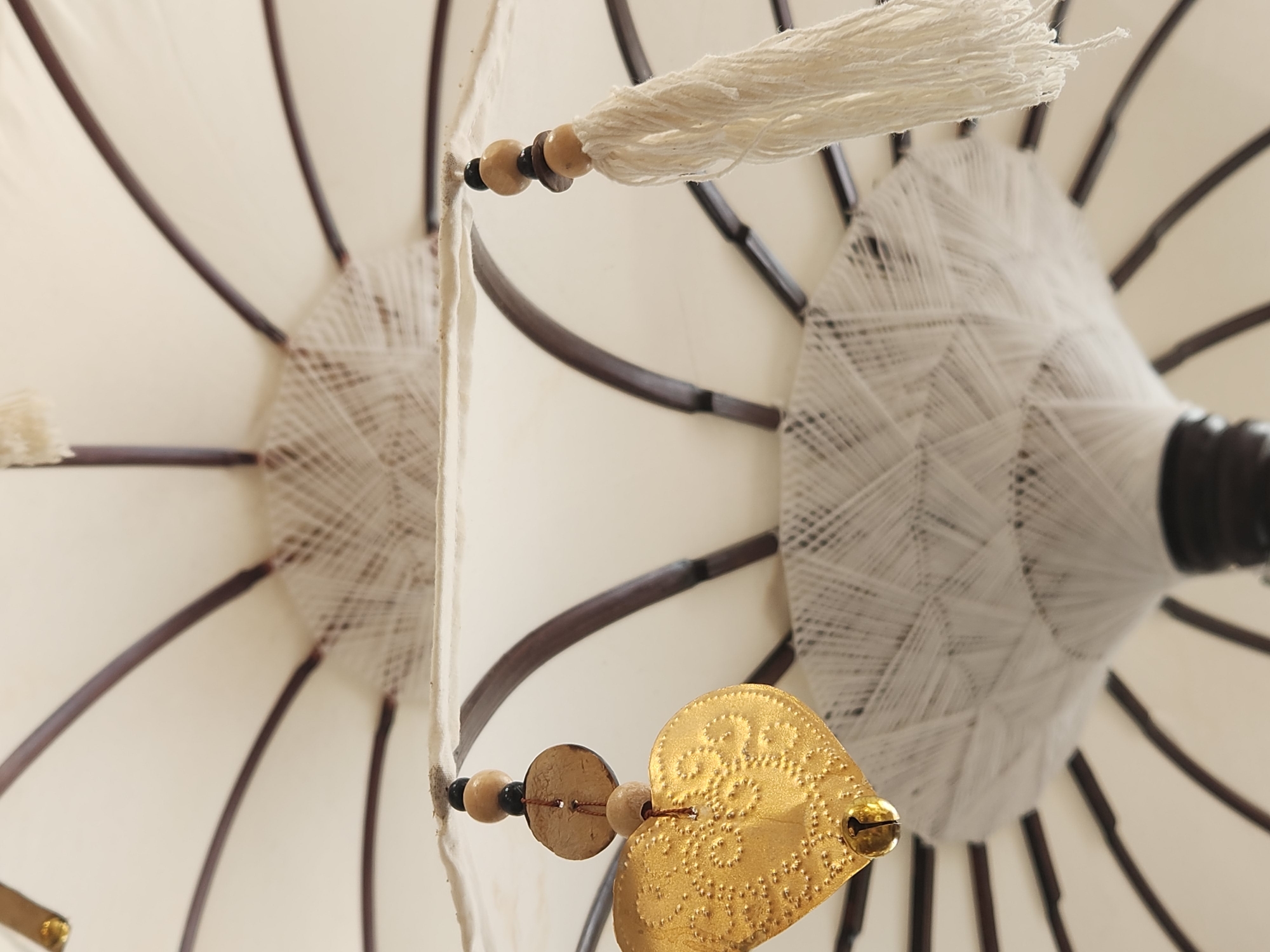 The image is a close-up, low-angle shot of the underside of a white umbrella, possibly a decorative or ceremonial one. The umbrella has dark brown ribs radiating from the center, and a white, woven fabric or string detailing in the lower portion. Two decorative elements hang from the edge of the umbrella. On the left, a golden, heart-shaped pendant with intricate embossed patterns and a small bell at the bottom hangs from a string adorned with wooden beads and a slice of what appears to be coconut shell. On the right, a white tassel hangs from a similar string of beads. The background is out of focus, showing more of the umbrella's structure. There are no people or animals visible in the image, and the environment suggests an indoor setting or a shaded outdoor area. The provided information does not contain details to determine its location in Maitland, United States of America.