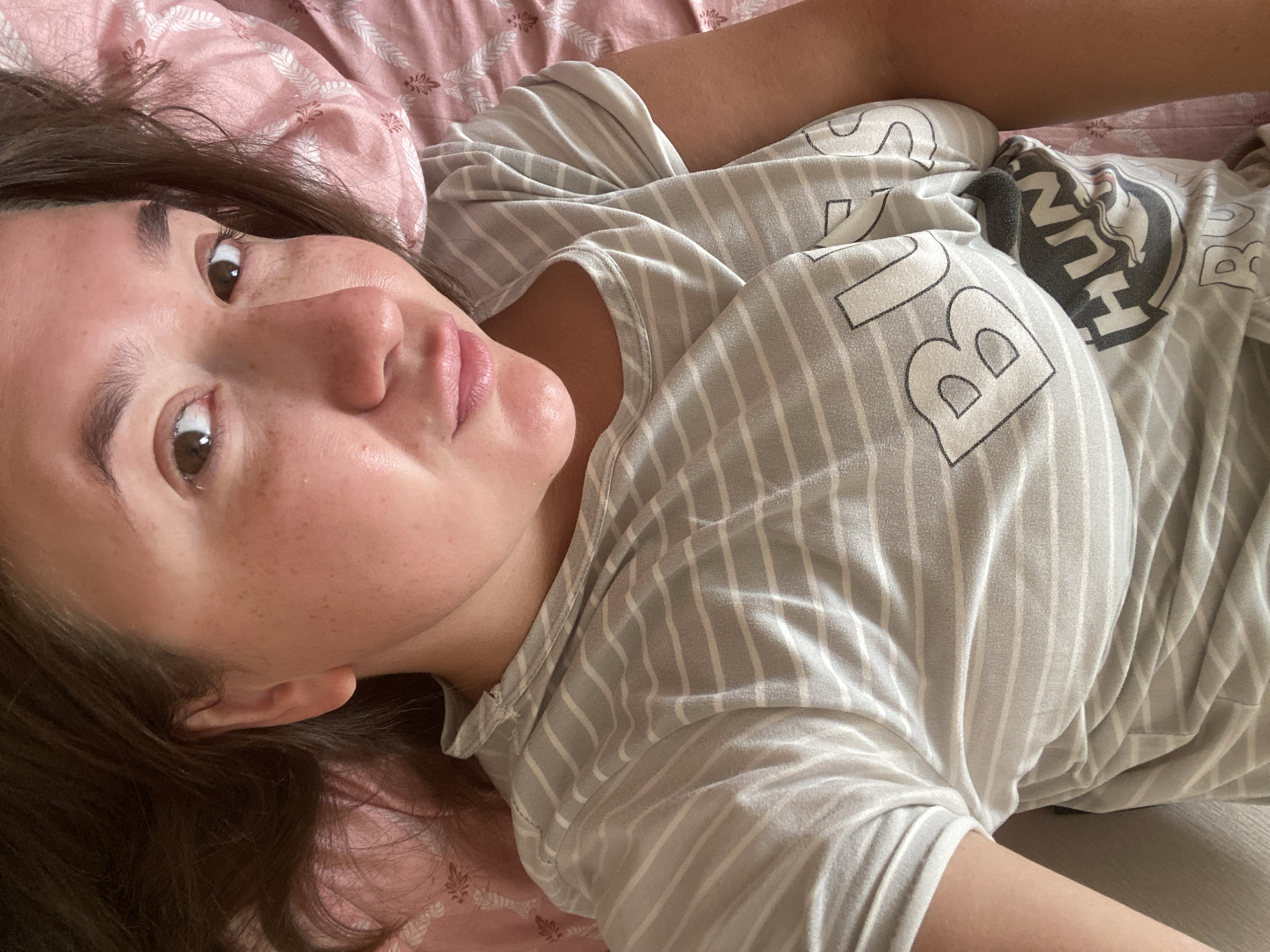 A woman is lying on her back on a pink and white floral patterned bed, facing upwards and looking at the camera. She has long, dark brown hair spread around her head. She is wearing a light gray t-shirt with thin horizontal white stripes. The t-shirt has text and graphics on it, including the word "BUS" partially visible on the chest, and a circular graphic with the word "HUNT" inside. Her skin has a warm tone and is visible with freckles on her cheeks and nose. Her lips are slightly parted. The background consists of the pink and white floral patterned bedsheets and some of her brown hair. The lighting appears to be natural and soft, creating some shine on her skin. The overall impression is of a casual, relaxed moment. The image was taken in Chaikovsky, Russia.