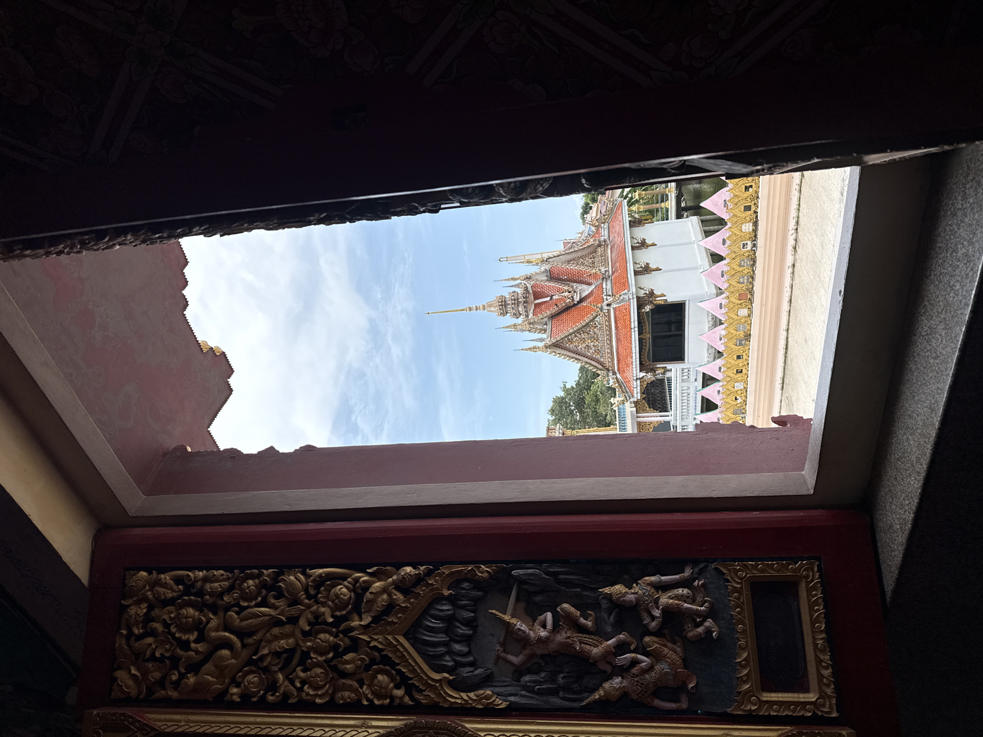 The image shows a view through a window or doorway of a building, likely a temple, in Ko Samui District, Thailand. The foreground features intricate golden carvings on what appears to be a wooden door or panel. These carvings depict figures in a scene with floral motifs.
The opening reveals a view of the exterior. Dominating the exterior view is a white building with a prominent, multi-tiered roof adorned with orange tiles and ornate golden spires. This structure, characteristic of Thai temple architecture, is surrounded by decorative golden fencing with triangular pink accents. In the background, lush green trees are visible, and the sky is a pale blue with scattered white clouds. The overall scene suggests a serene and culturally rich environment.