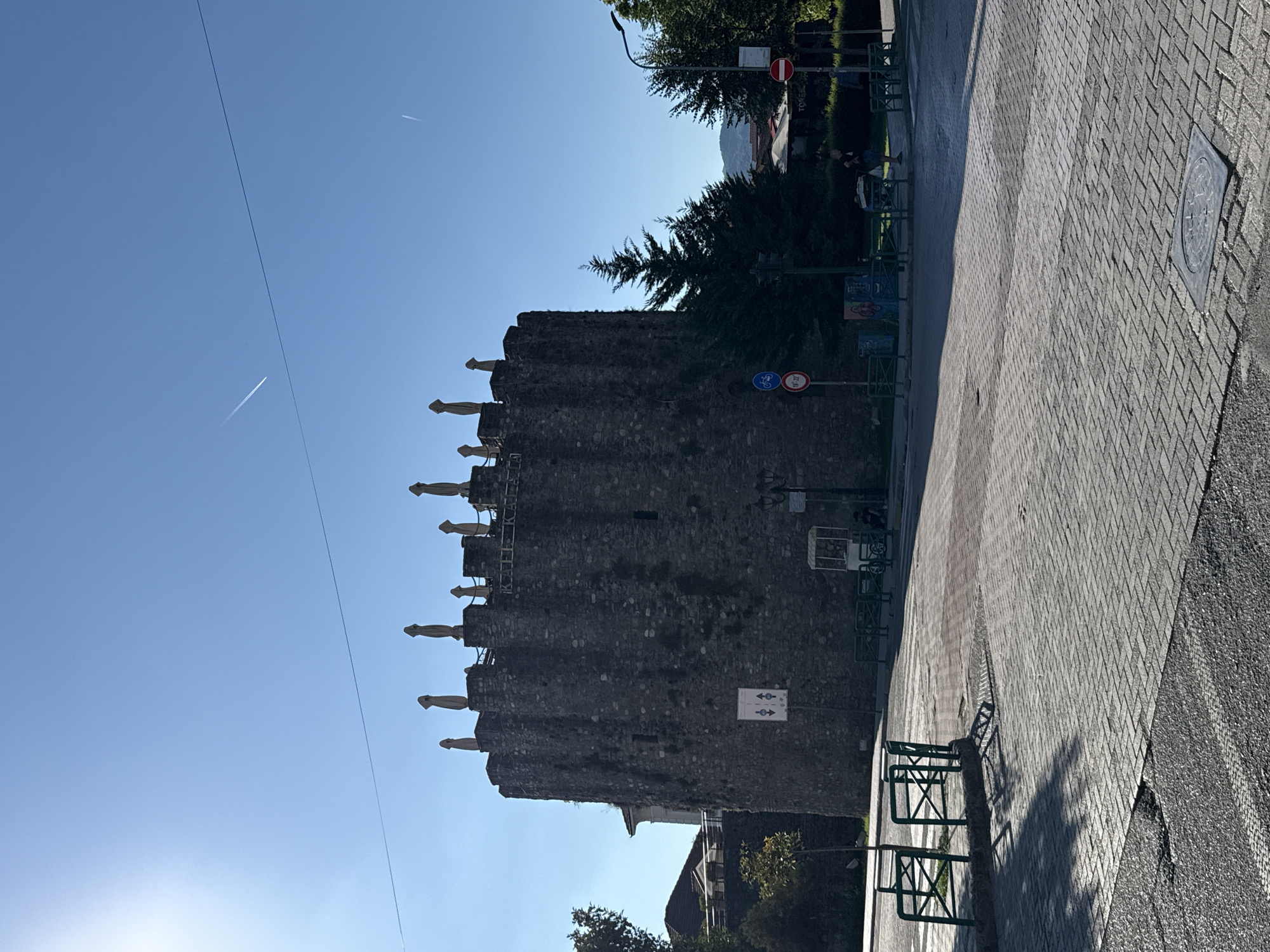 The image depicts a sunlit outdoor scene in Elbasan, Albania, featuring a historical stone tower as the central subject. The tower, with its crenellated top, appears ancient and robust, adorned with lighter-colored awnings or coverings along its battlements. Adjacent to the tower is a paved walkway made of interlocking grey bricks, transitioning to a darker, asphalt-like surface on the right. Green metal bike racks are visible along the walkway, suggesting a public space.
The sky is a clear, vibrant blue, with two faint contrails from airplanes indicating the presence of air traffic. A thin, dark line, possibly a cable or wire, stretches diagonally across the sky, intersecting with the tower. In the background, lush green trees and the corner of a building with a red-tiled roof are visible, hinting at an urban or semi-urban environment. Sunlight casts distinct shadows from the bike racks onto the pavement, suggesting it is either morning or late afternoon.
Several signs are present: one circular red and white sign, likely a prohibition, is partially visible at the top; below it, a circular sign with a bicycle and a number "20-22" indicates a bike lane or parking area. Another sign features blue arrows pointing in opposite directions, possibly indicating a one-way street or traffic flow. A small white sign with two figures and arrows is also discernible on the tower. No people are clearly visible in the image, though the presence of bike racks and public paving suggests it is a place frequented by people. The overall impression is one of a bright, clear day in a location that blends historical architecture with modern amenities.