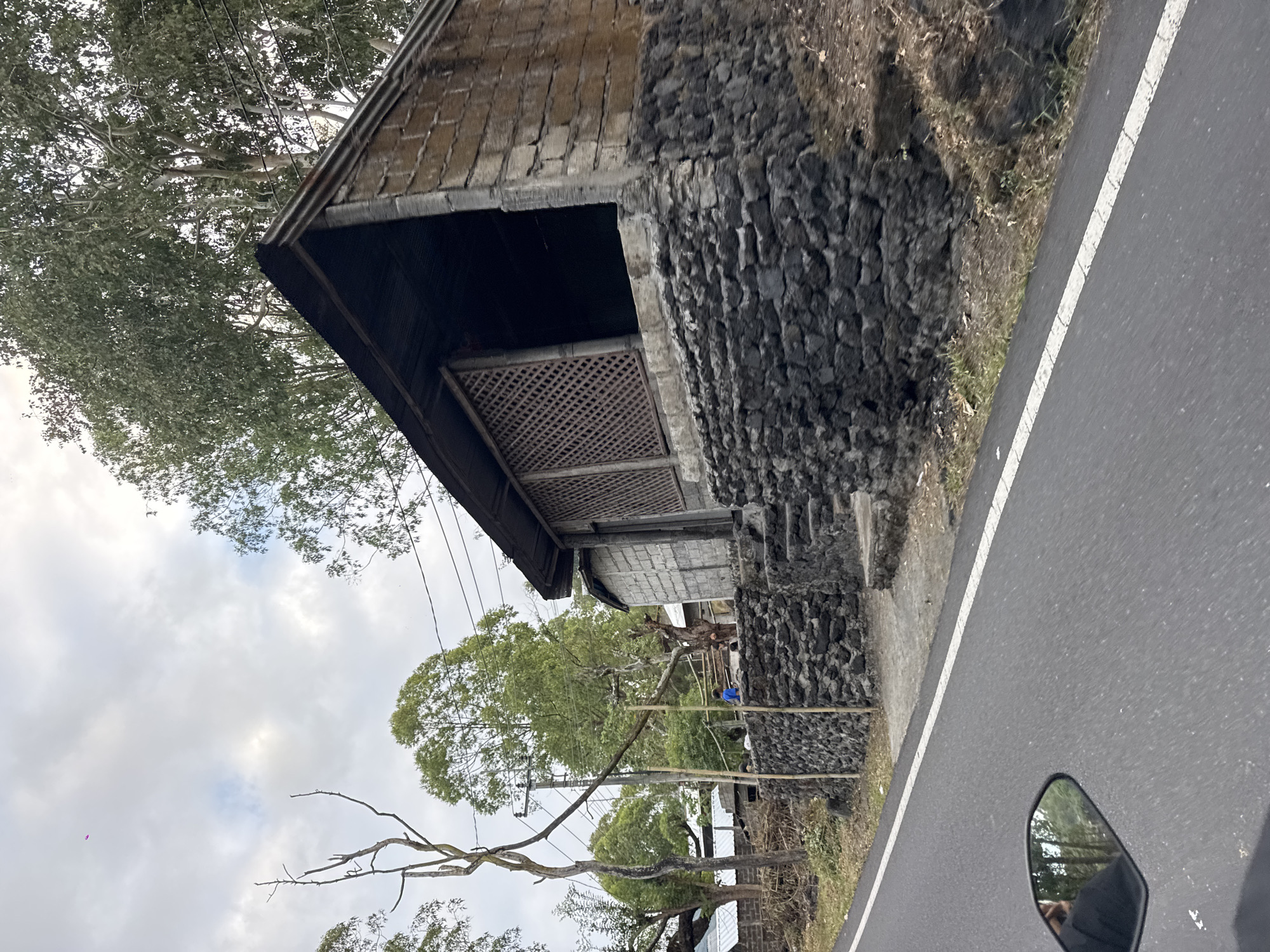 This image depicts a roadside scene in Kintamani, Indonesia. A simple, open-air structure constructed from stone and concrete block stands on the right side of the frame, elevated by a dark stone foundation. The structure has a corrugated metal roof and a lattice-like screen on one side. Stone steps lead up to its entrance. In the background, lush green trees and a utility pole with wires are visible against a cloudy sky. A paved road with a white dashed line runs along the left side of the image, and a motorcycle's rearview mirror, reflecting trees and a person, is visible in the lower left corner. Several smaller structures with metal roofs are visible further in the background to the left of the main building. A young child in a blue shirt is visible standing on the stone wall in front of the main building, next to a person.