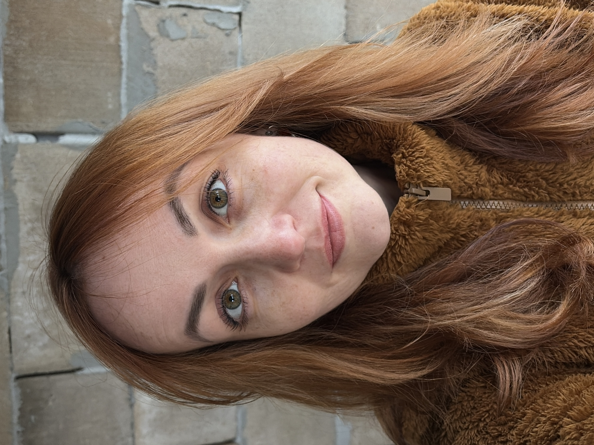 A close-up portrait shows a young woman with long, flowing reddish-brown hair. Her head is tilted, and she is looking directly at the camera with a gentle, slight smile. Her eyes are a striking green, framed by well-defined eyebrows and dark eyelashes enhanced with mascara. Freckles are visible across her nose and cheeks. Her lips are a natural, soft pinkish-brown.
She is wearing a cozy, brown, fuzzy jacket or coat with a visible zipper and zipper pull on the right side of the frame. The material of her outerwear appears soft and textured. The lighting is soft and even, highlighting her features without harsh shadows, suggesting an overcast day or well-diffused natural light. In the background, a light-colored brick or stone wall is visible, indicating an outdoor or semi-outdoor setting. The wall consists of rectangular blocks with mortar lines. No specific activity beyond posing for the photograph is depicted. There is no visible text in the image.