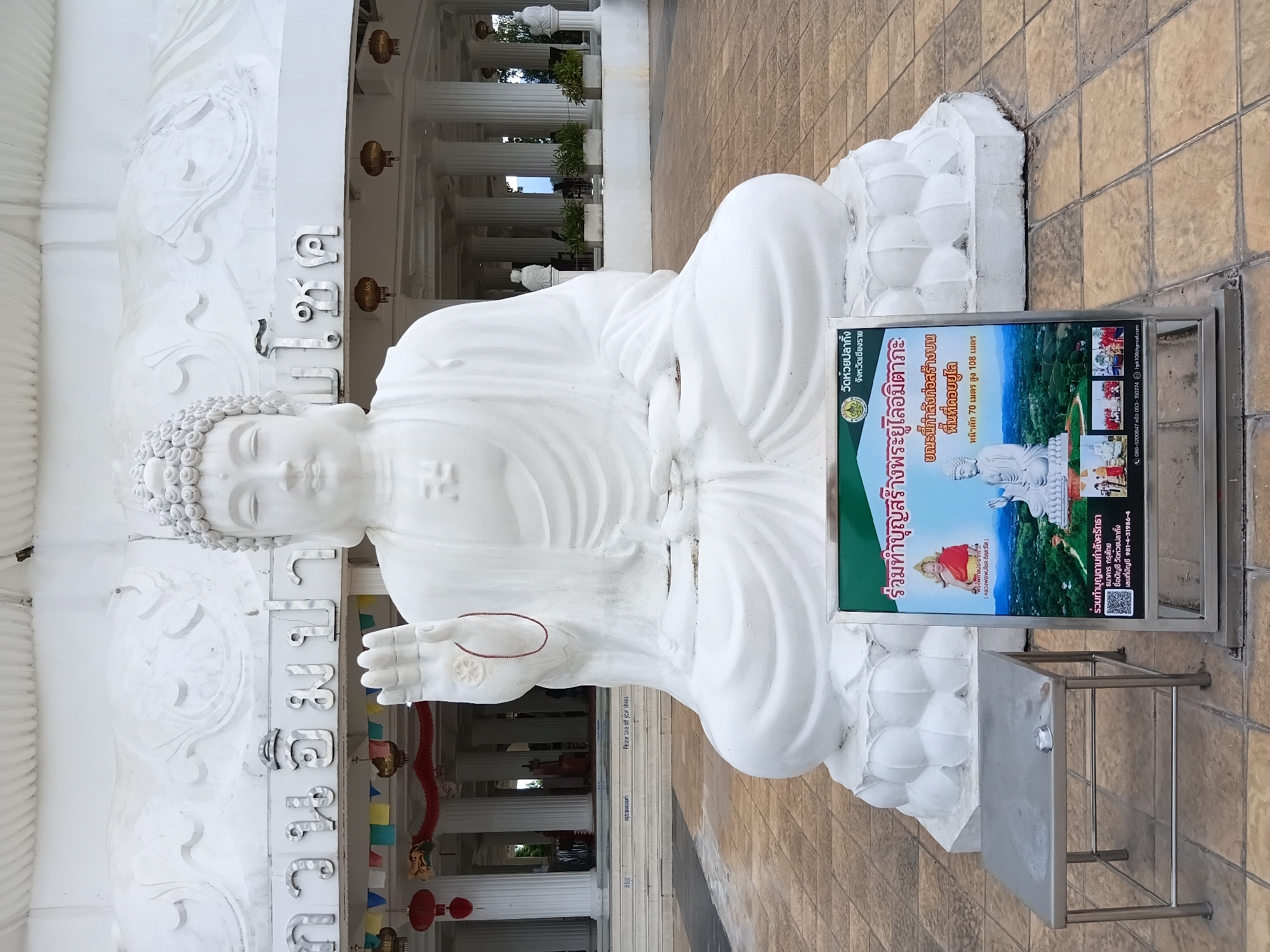 A large, white, seated Buddha statue is positioned in the foreground, facing the viewer. The statue is on a lotus pedestal, and the Buddha's right hand is raised, palm outward, with a small red string tied around the wrist. Behind the statue is a white building with elaborate carvings and decorative lanterns. In front of the Buddha statue, on the ground, is a metal table with an ashtray and a sign with images and Thai text. The ground is paved with square tiles. The overall scene suggests a temple or religious site.