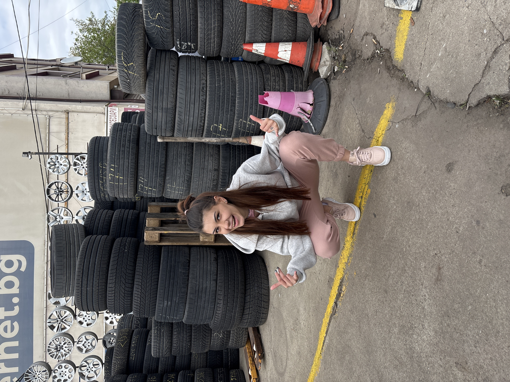 A young woman with long brown hair stands in a low squat next to a yellow line on a concrete surface, positioned next to a large stack of tires and a display of car rims. She wears a light gray hoodie and pink sweatpants, with matching pink sneakers. She is gesturing with both hands, pointing her index fingers toward the ground and smiling at the camera. Orange and white traffic cones are visible adjacent to the tires. A sign with the text "internet.bg" can be seen among the car rims. The setting appears to be a tire shop or automotive service area. The city is Sofia, Bulgaria.